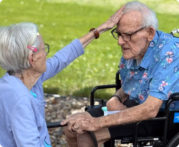 Elderly woman gently touches the forehead of an elderly man sitting in a wheelchair outdoors.