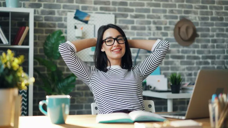 A young woman in her home office, leaning back in her chair in front of her laptop while working.