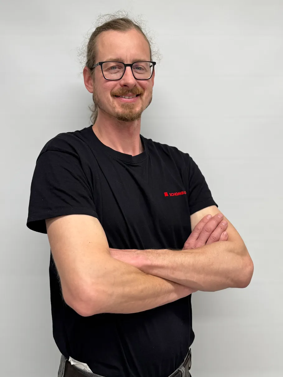 Smiling man with glasses and a beard wearing a black Schombu t-shirt, standing with arms crossed.