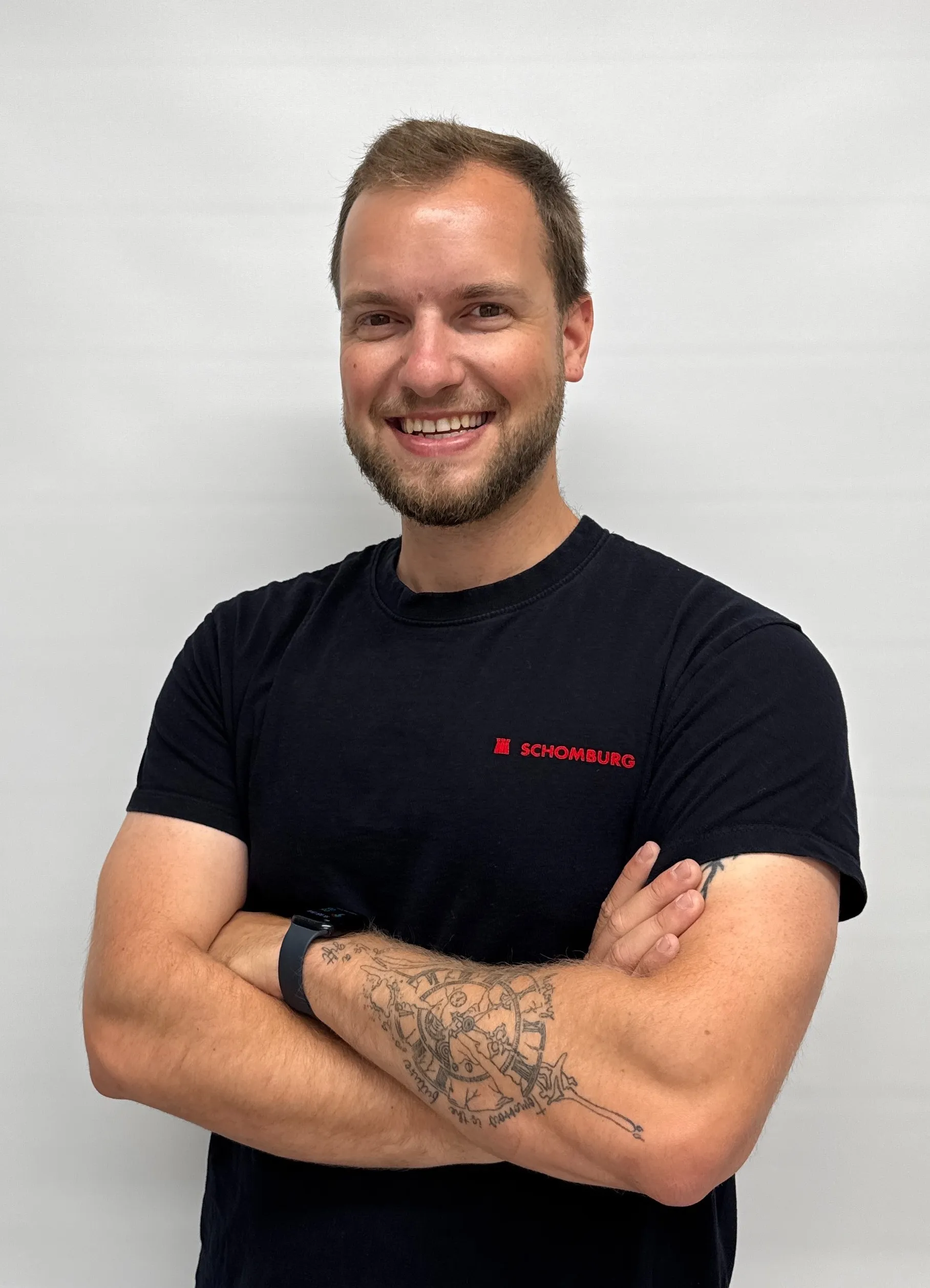 Smiling man with short hair and beard wearing a black Schomburg T-shirt and a smartwatch, with a detailed clock tattoo on his forearm.