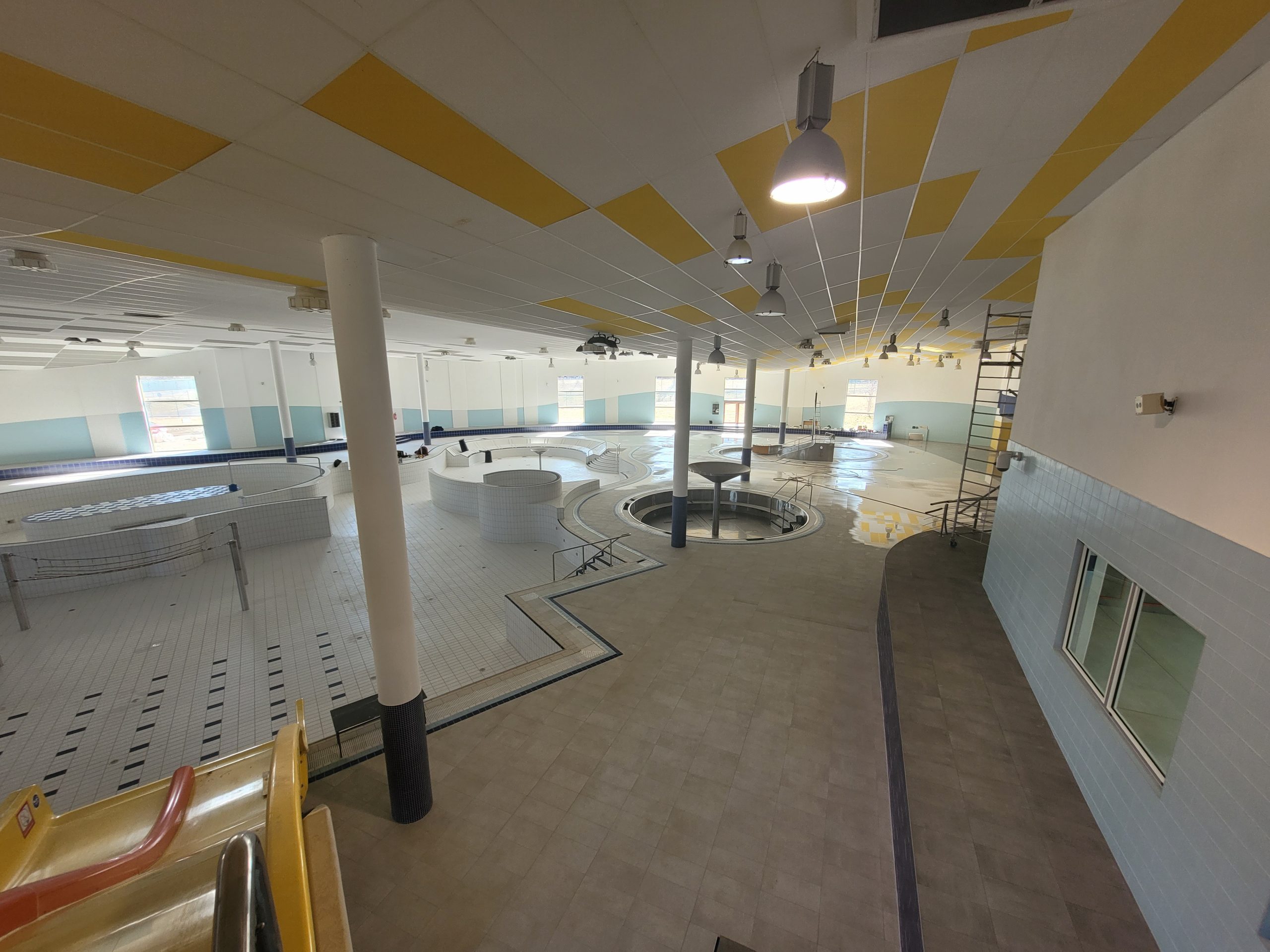 Empty indoor swimming pool area with white tiled floors, circular pools, yellow ceiling panels, and hanging lights.