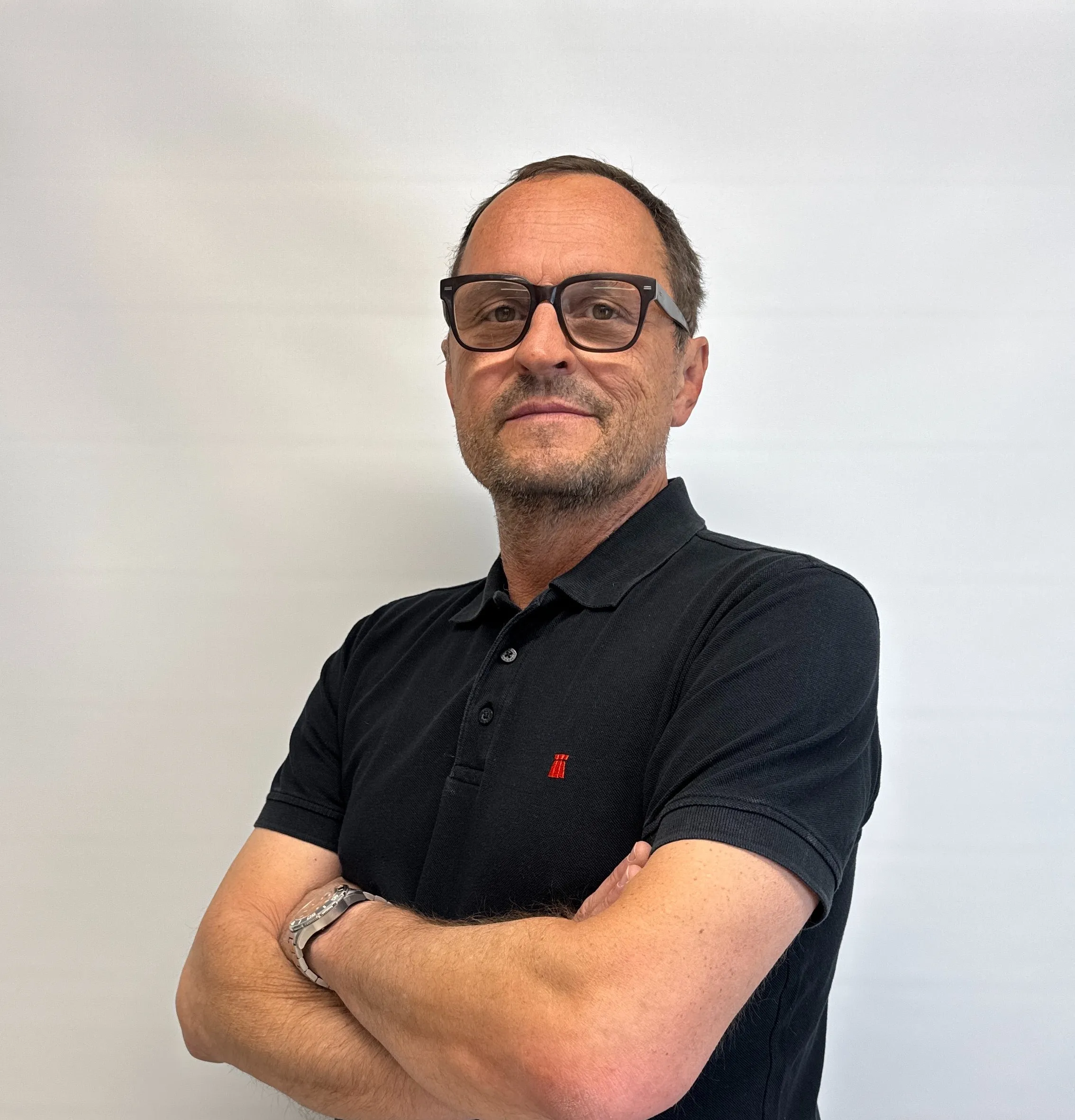 Man with glasses and a short beard wearing a black polo shirt with folded arms, standing against a plain light background.