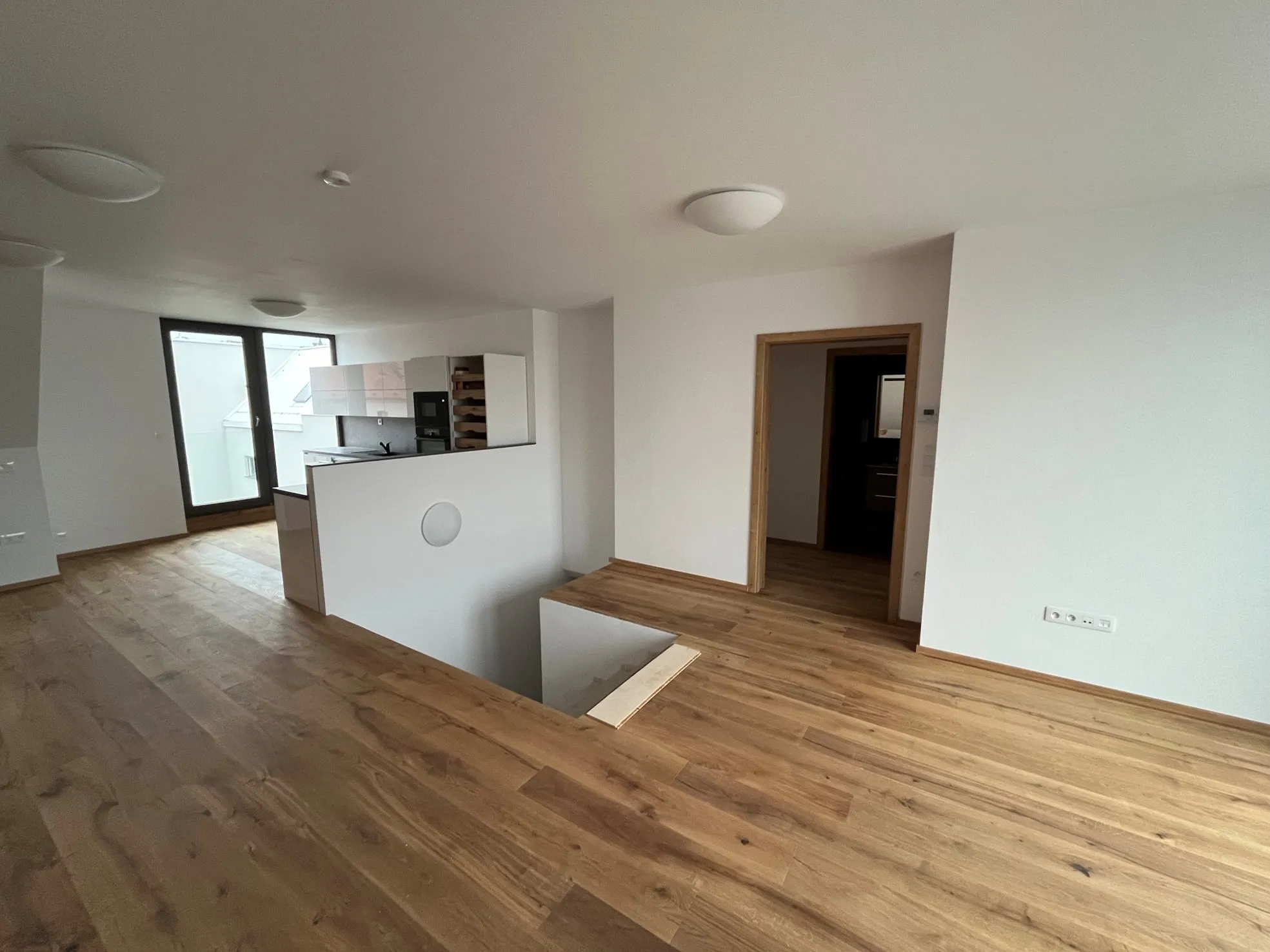 Modern empty apartment interior with wooden flooring, white walls, a small kitchen area, and a staircase leading downstairs.