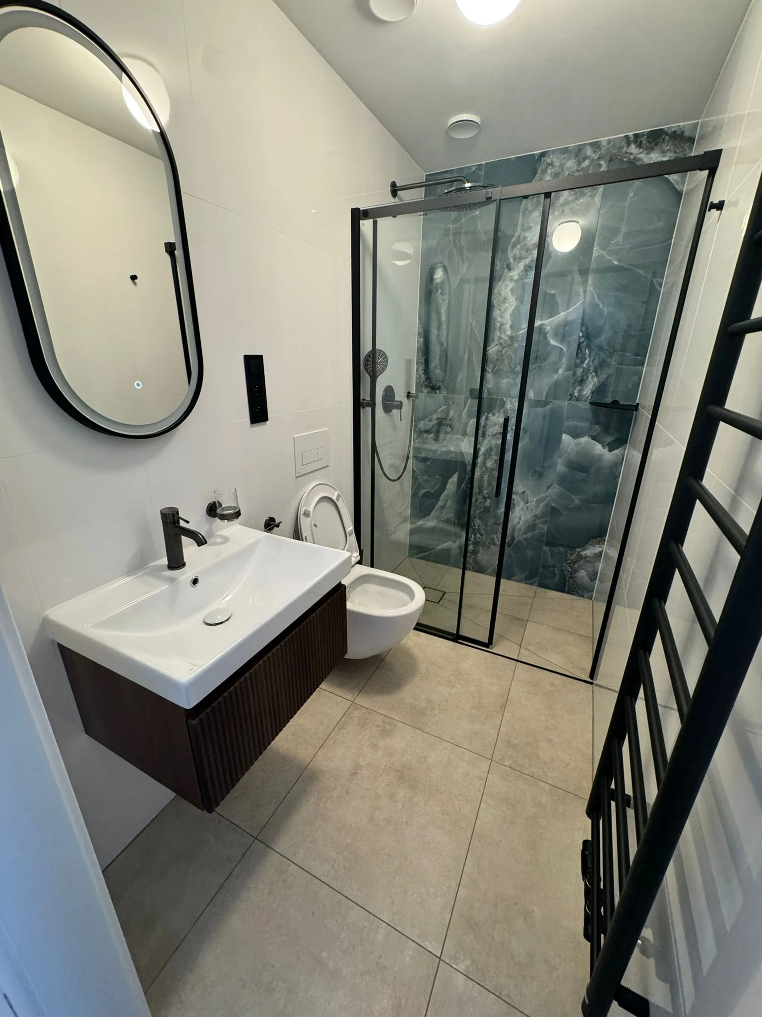 Modern bathroom with white sink, wall-mounted toilet, glass shower enclosure with blue marble wall, and black towel radiator.