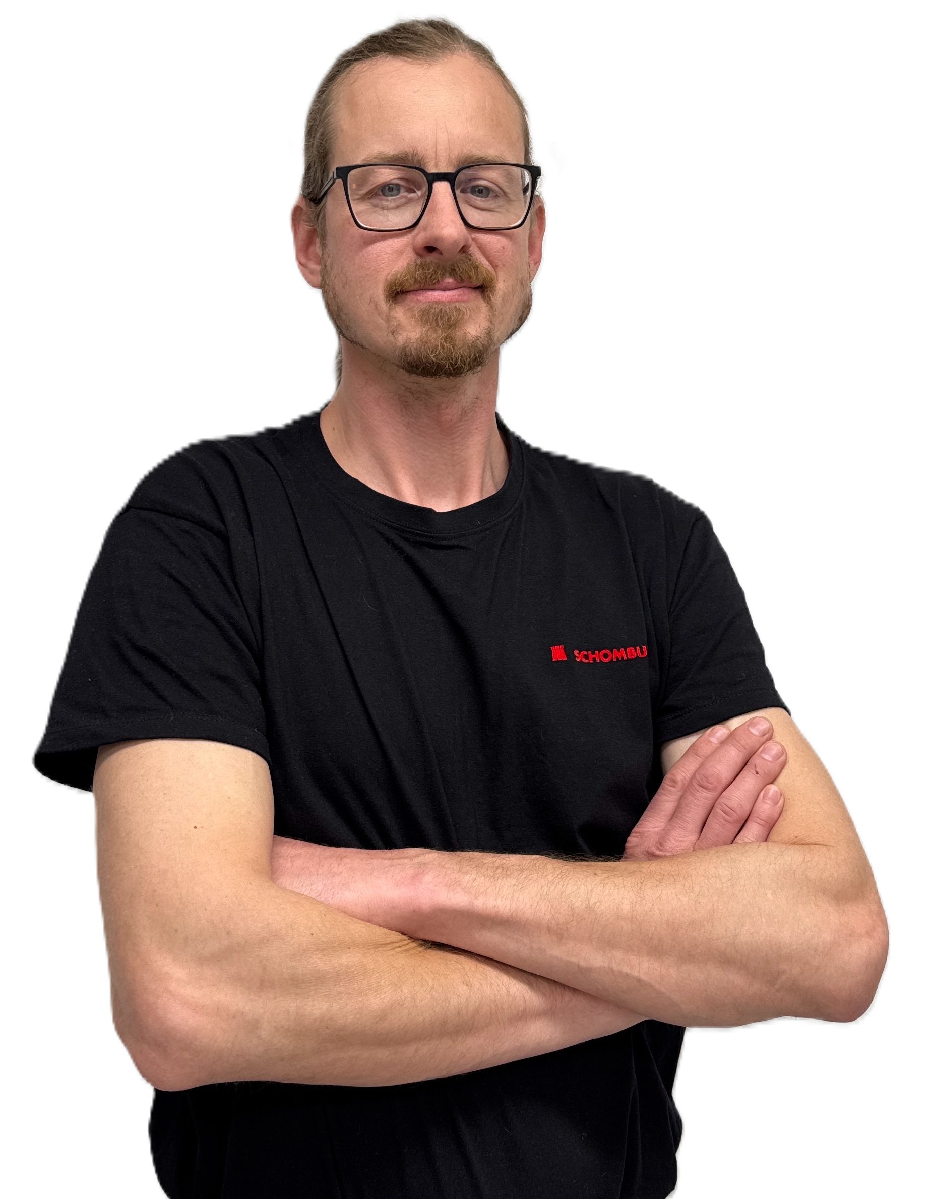 Smiling man with glasses and a beard wearing a black Schombu t-shirt, standing with arms crossed.