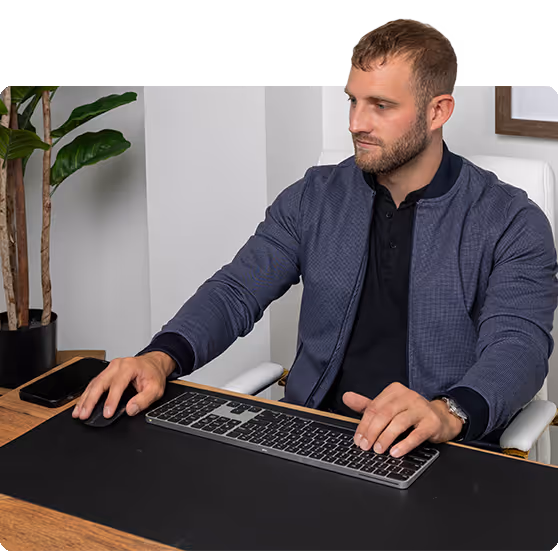 Man in blue jacket working on computer at wooden desk with plant