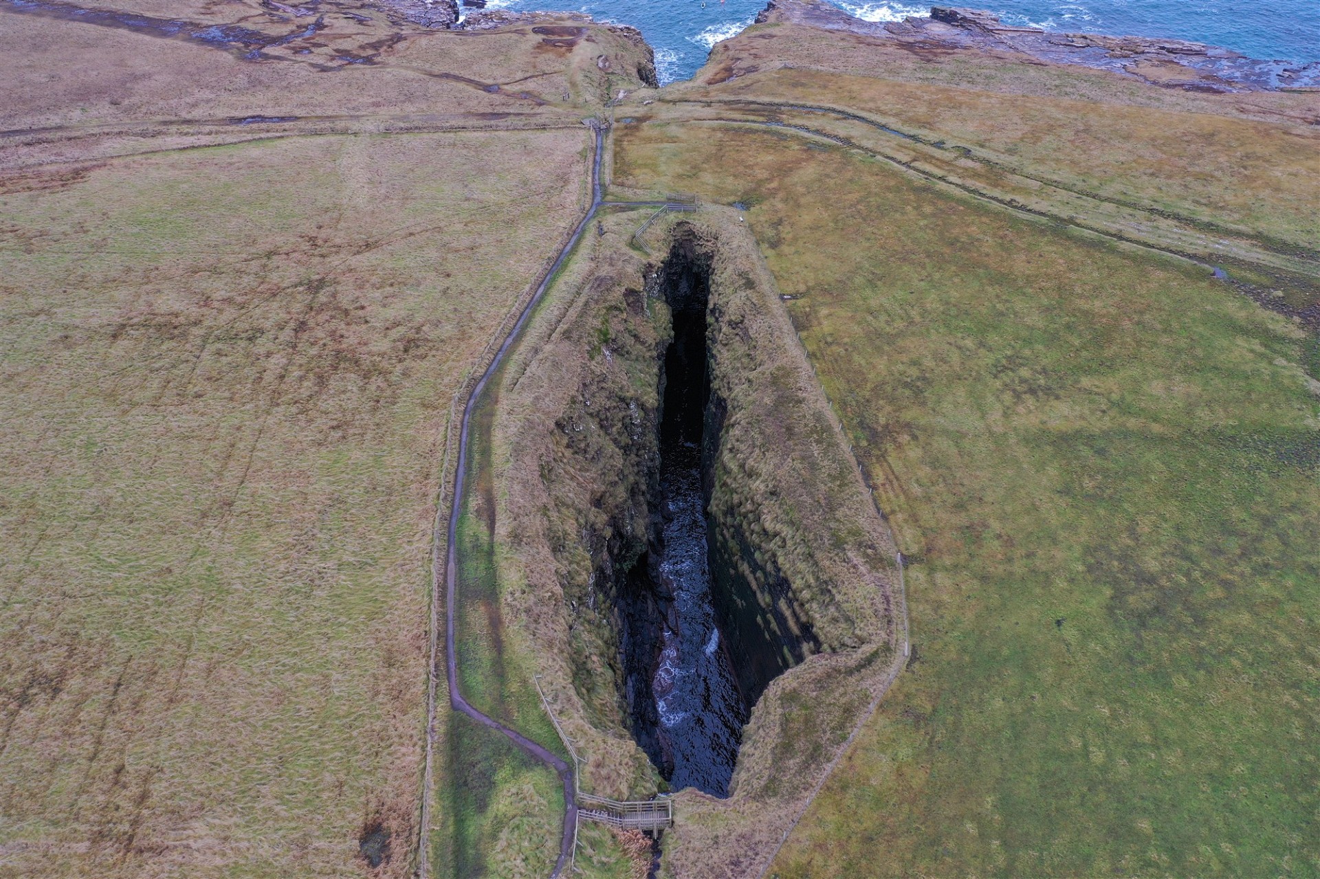 Aerial view of a narrow, deep coastal chasm surrounded by grassy fields with a footpath and wooden viewing platforms on either end.
