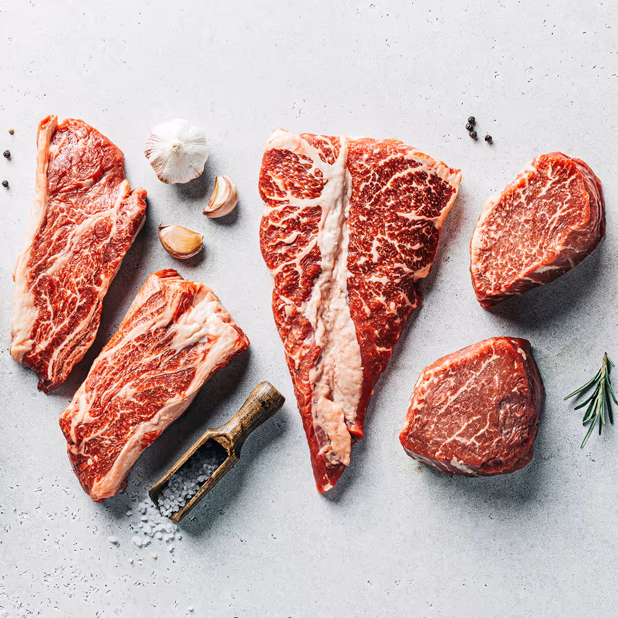 Assorted raw beef cuts with visible marbling, arranged on a light surface with garlic cloves, coarse salt, peppercorns, and a sprig of rosemary.
