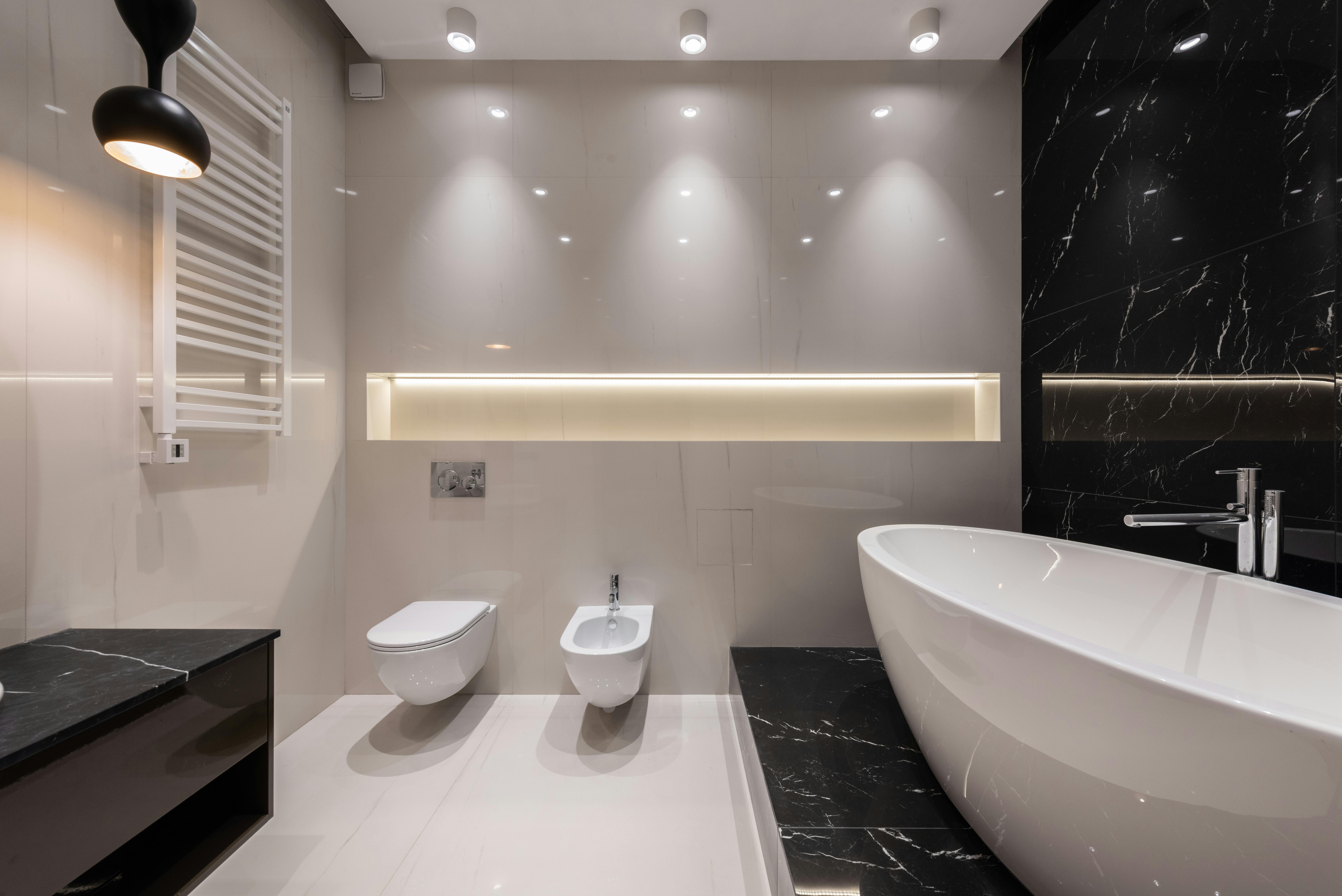 Modern bathroom with a freestanding white bathtub on black marble platform, wall-mounted toilet, bidet, and a black pendant light.