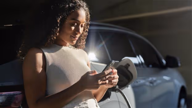 Frau lehnt an E-Auto beim Laden und schaut auf ihr Smartphone in sonniger Parkgarage.