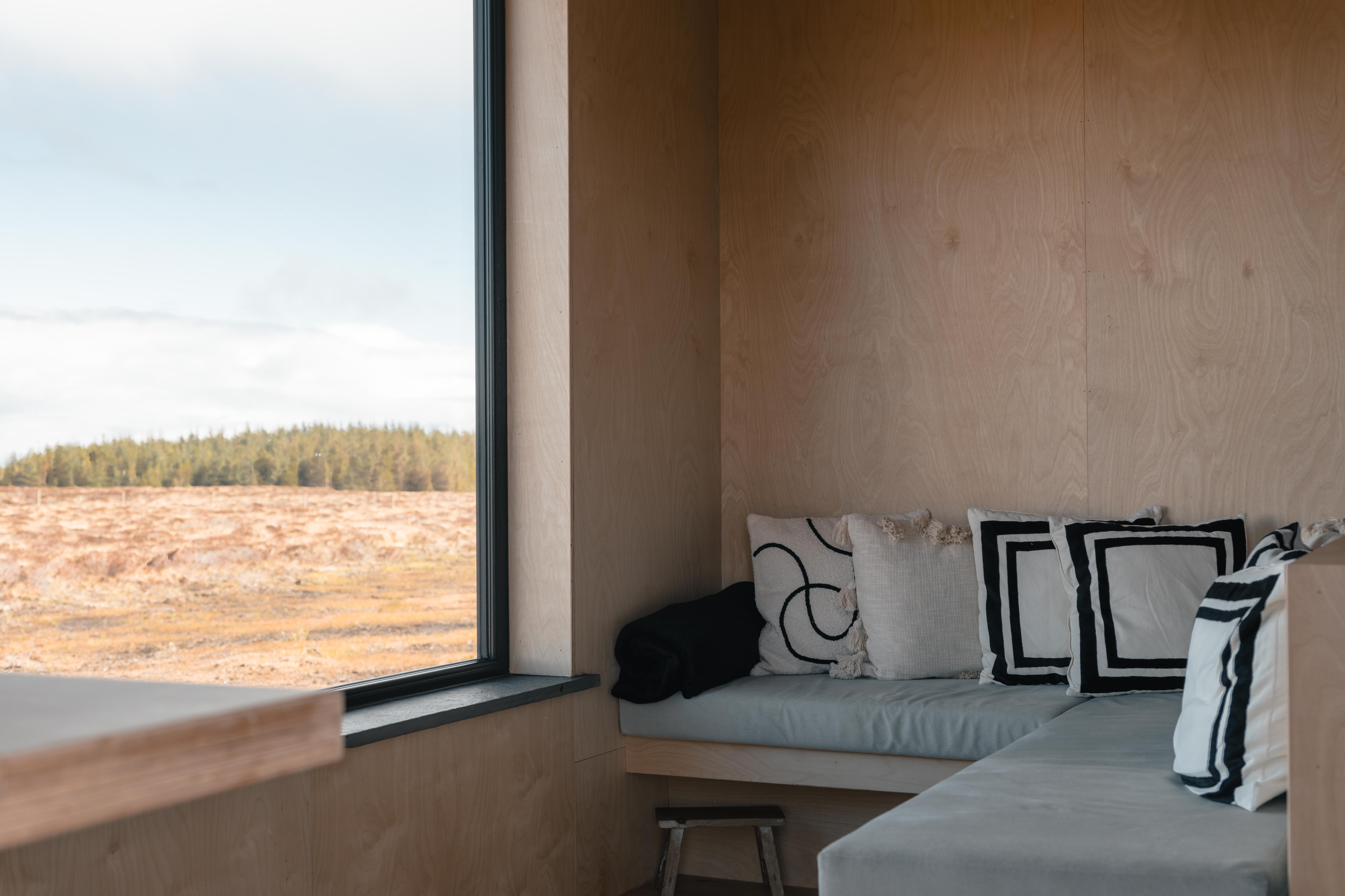 Cozy wooden room with a large window overlooking a rainy landscape and a cushioned bench with decorative pillows.