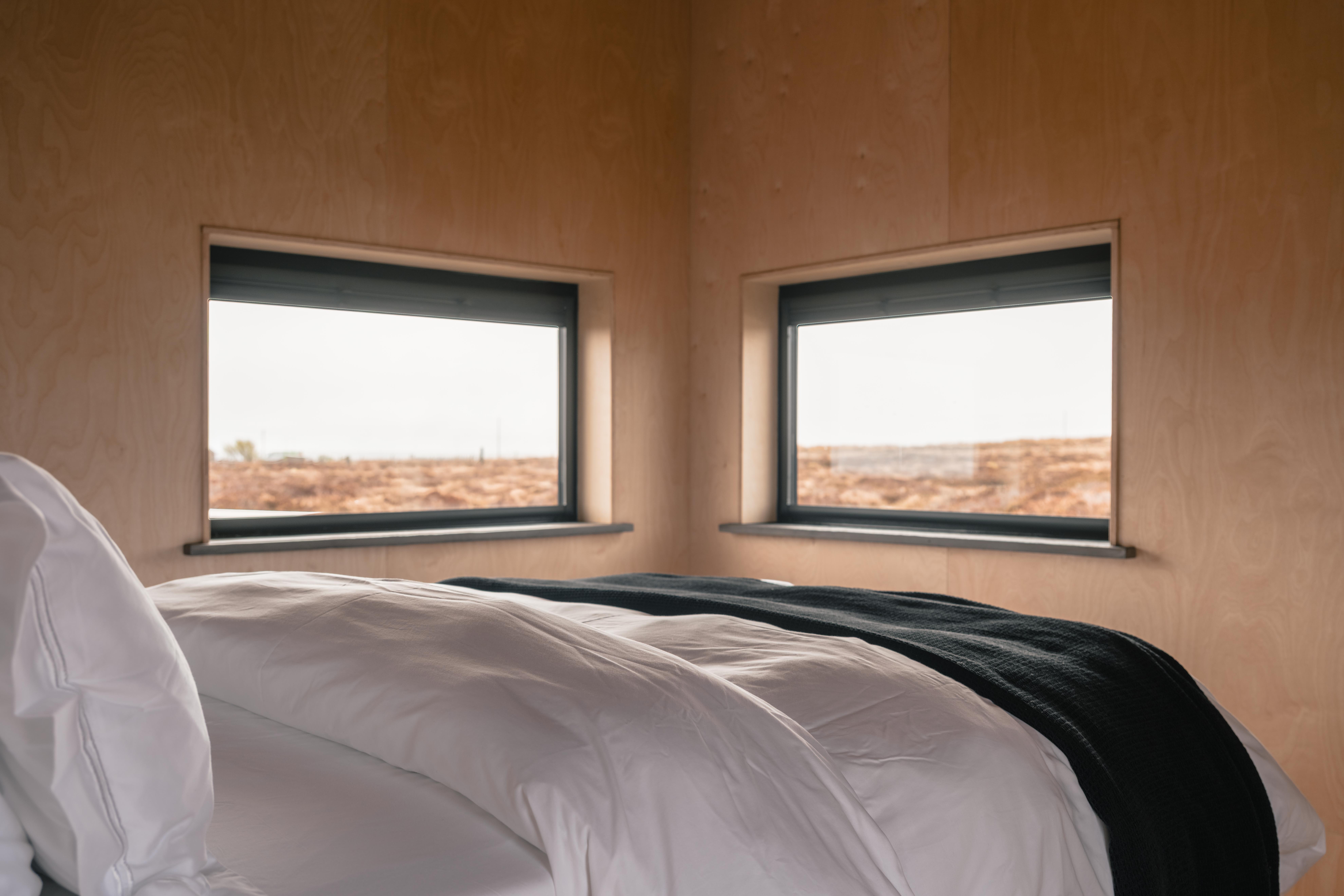 Cozy bedroom with a neatly made bed, wooden walls, two windows overlooking a natural landscape, a wall-mounted TV, and folded towels on the bed.