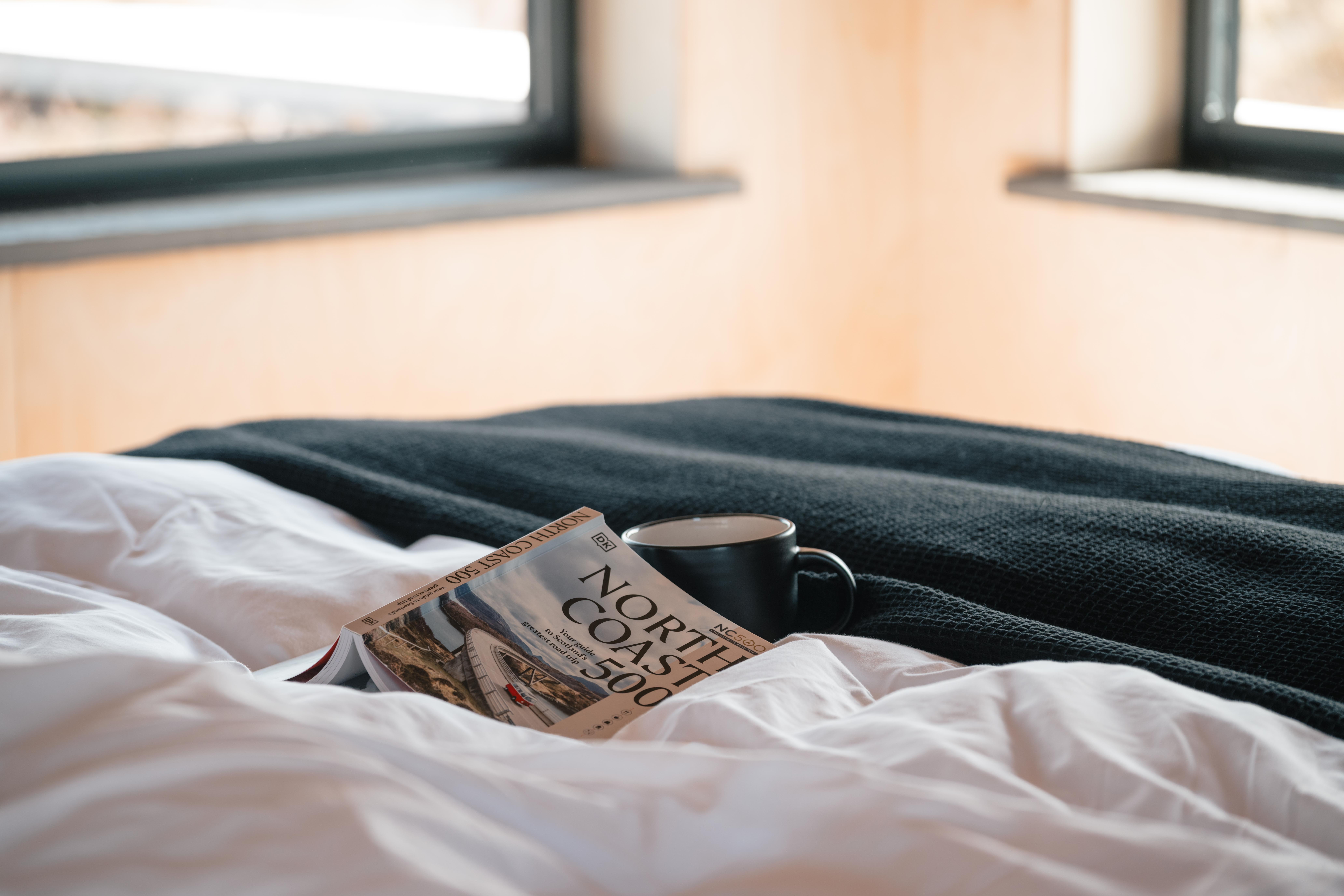 Minimalist bedroom with wooden walls and floor, a bed with white linens, beige throw, and folded blankets, two small wall windows overlooking a landscape.