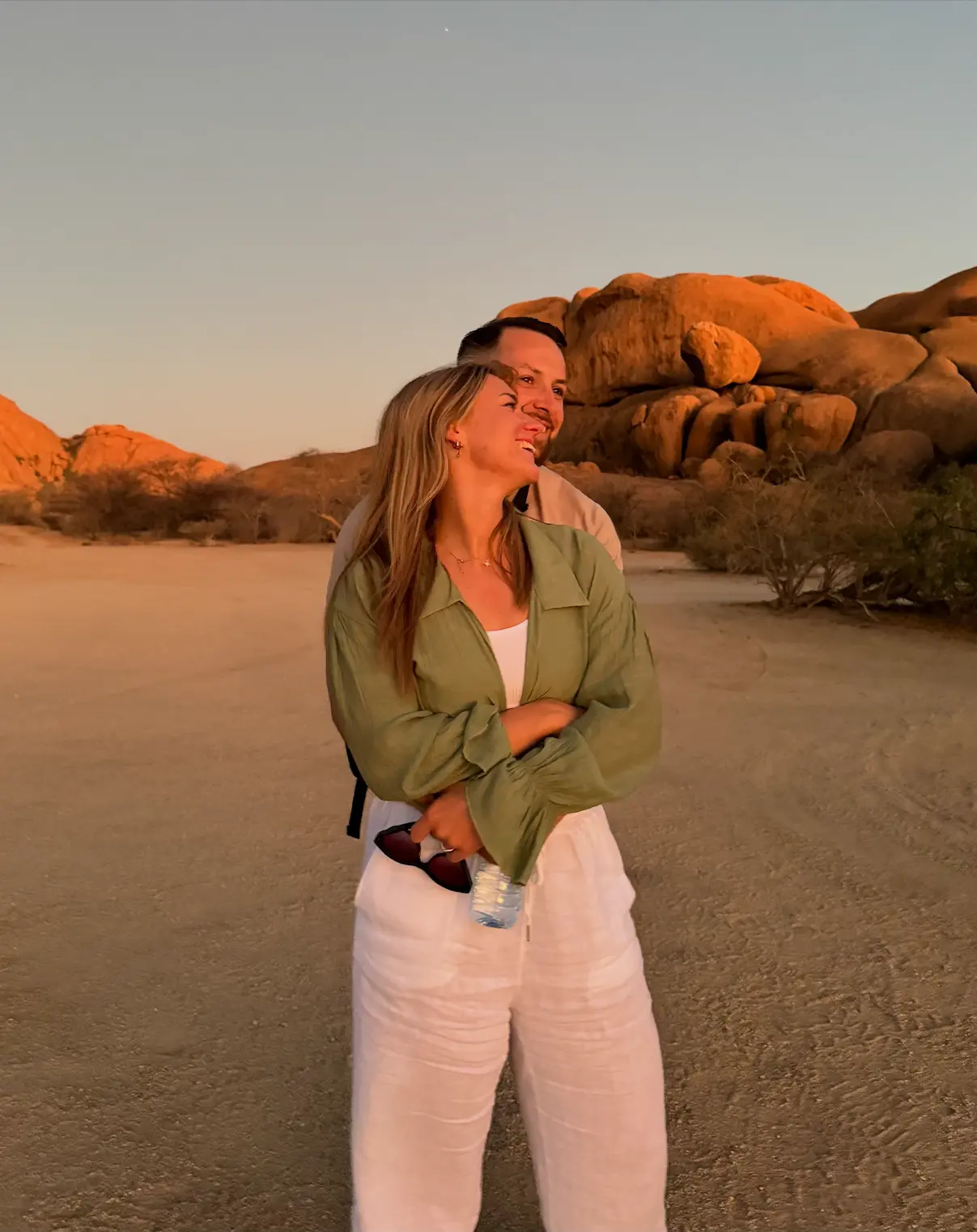 Paar umarmt sich in einer Wüstenlandschaft mit Felsen im Hintergrund bei Sonnenuntergang.