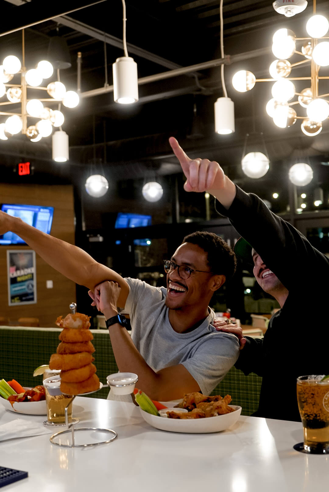 Two friends cheer and laugh over food and drinks at a lively restaurant.