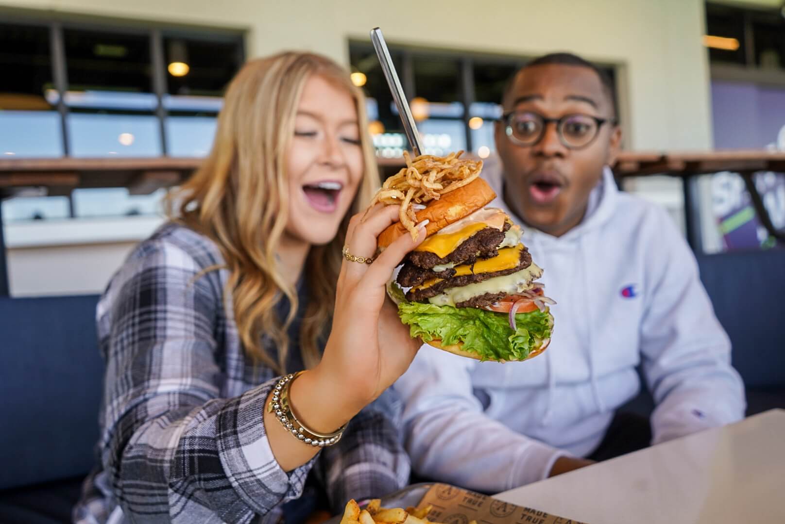 Two friends react excitedly to a giant stacked burger at a restaurant.