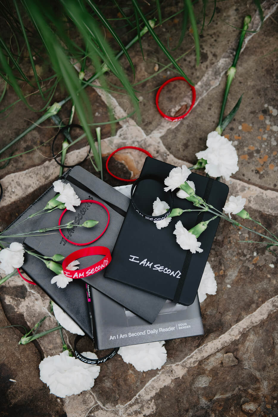 Notebooks and white flowers arranged on a stone surface, surrounded by red and black "I AM SECOND" wristbands.