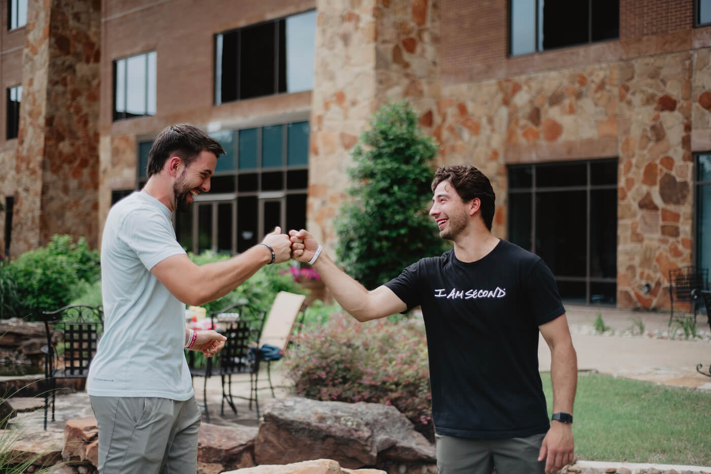 Two men smiling and doing a fist bump outside a building, one wearing an "I AM SECOND" shirt.