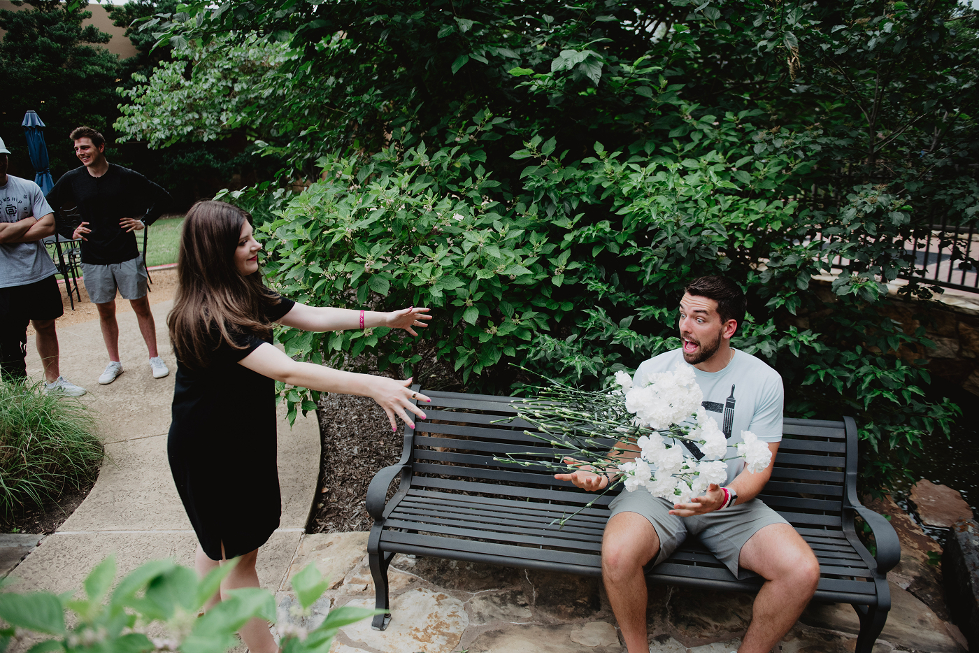 Woman playfully reaching for man holding white flowers on a bench, with friends watching nearby.