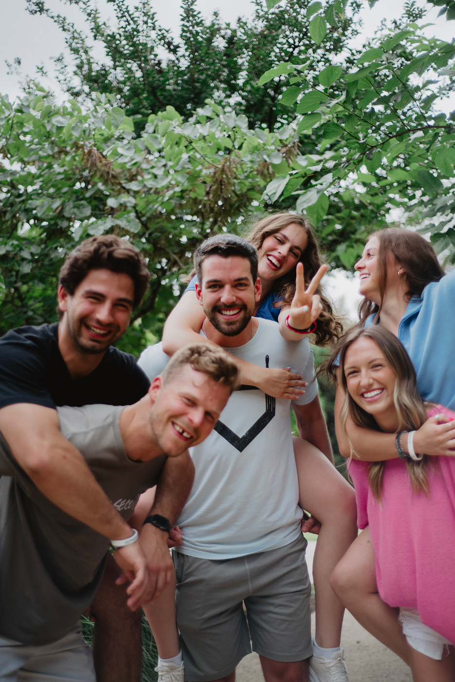 A group of smiling young adults posing playfully outdoors, some giving piggyback rides and wearing "I AM SECOND" wristbands.