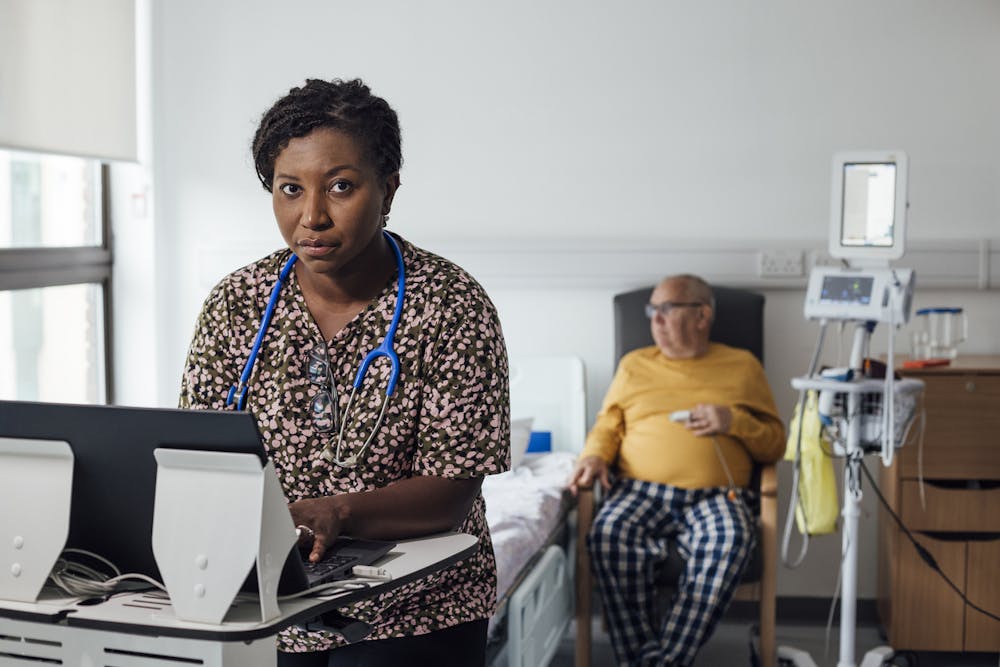 Nurse looks towards the camera as an elderly patient waits to be treated in the background.