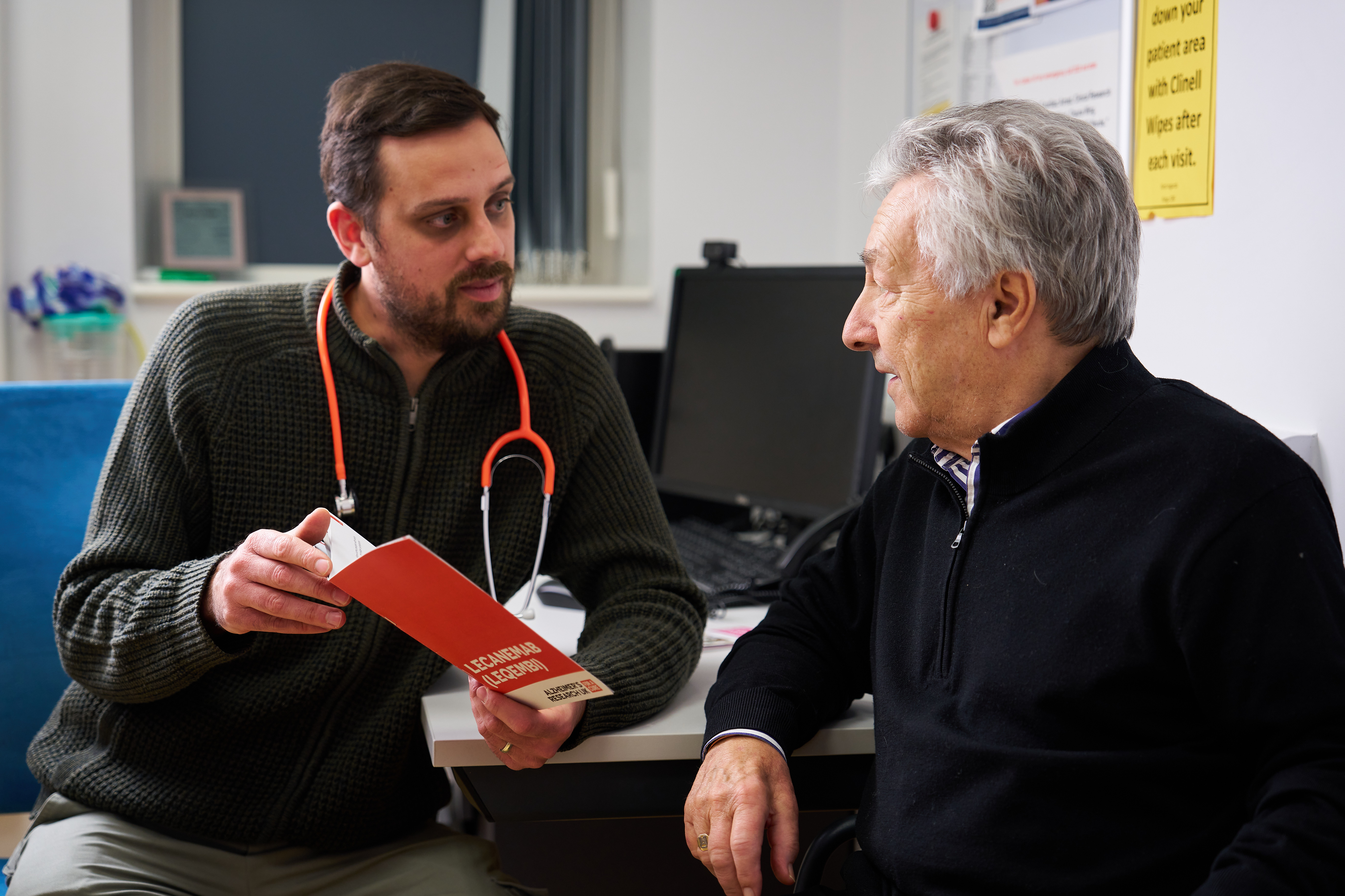 A GP speaking with one of his patients in the GP surgery while showing them the contents of an Alzheimer's Research UK brochure.