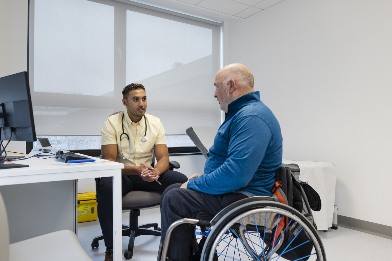 A male GP sits next to a desk facing a man in a wheelchair in the office of a GP surgery.