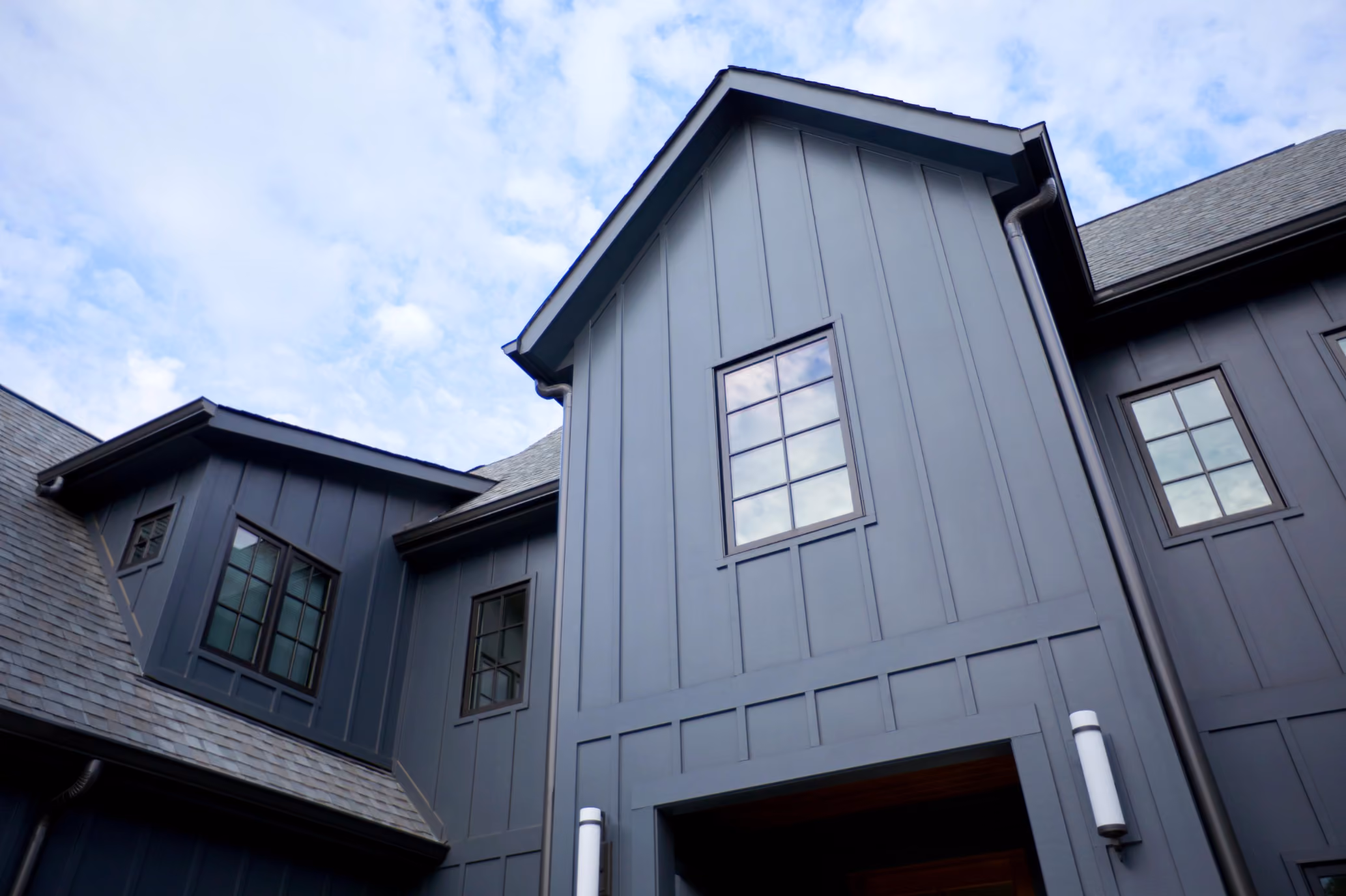 Exterior view looking up at a modern dark gray house with multiple windows and a sloped roof under a partly cloudy blue sky.