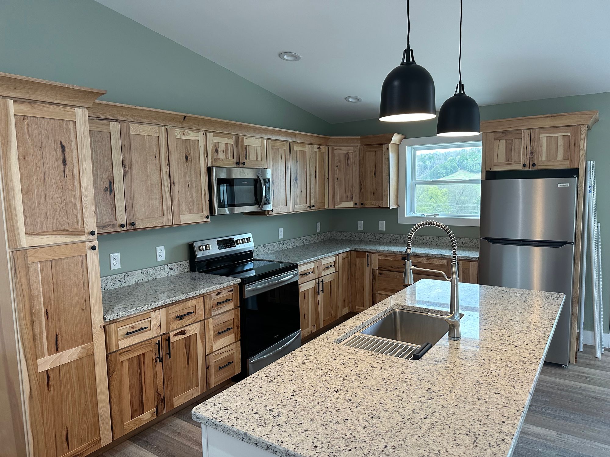 Modern kitchen with natural wood cabinets, granite countertops, stainless steel appliances including refrigerator, stove, and microwave, and a large island featuring a stainless steel sink with a high-arc pull-down faucet and two black pendant lights overhead, with a window showing greenery outside.