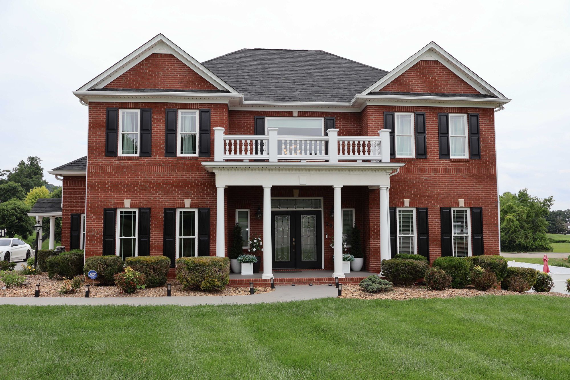 Two-story red brick house with a dark gray shingled roof, white balcony railing above the entrance, black shutters on the windows, white columns supporting a covered porch, and neatly trimmed bushes and green lawn in front.