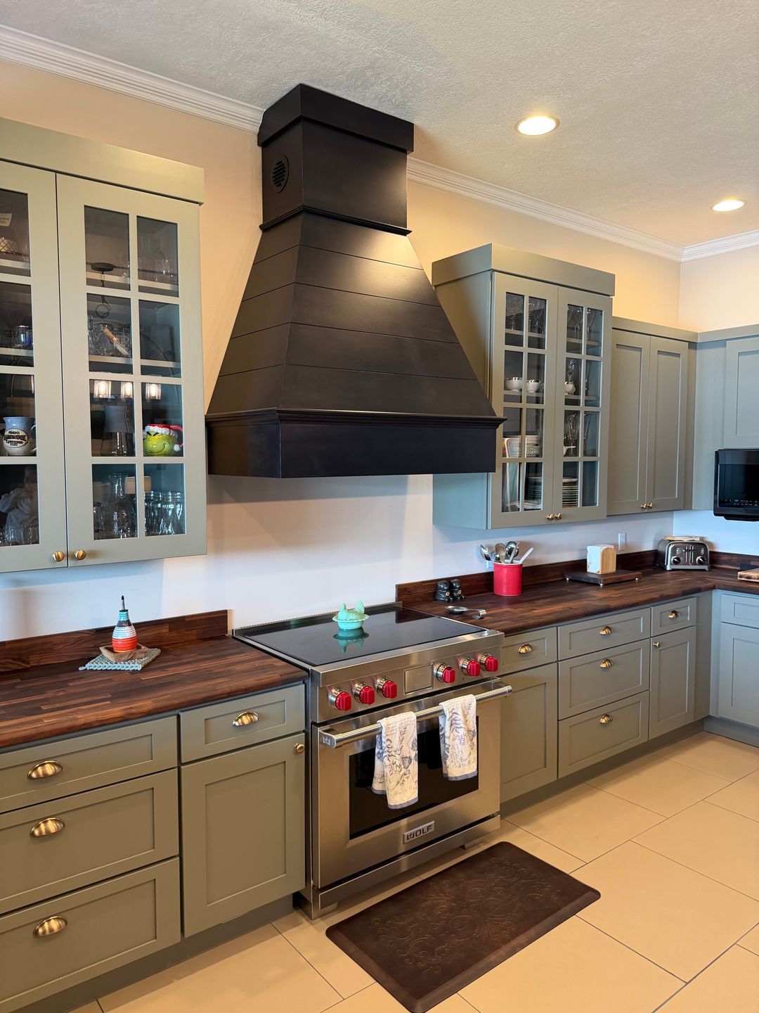 Modern kitchen with sage green cabinets, wooden countertops, stainless steel Wolf stove with red knobs, black angled range hood, and beige tile floor with a dark floor mat in front of the stove.