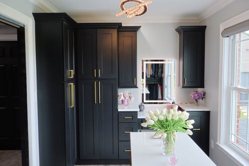 A modern closet with black cabinets featuring gold handles, white countertops, a lighted mirror, and vases with white tulips and purple flowers, next to a window.