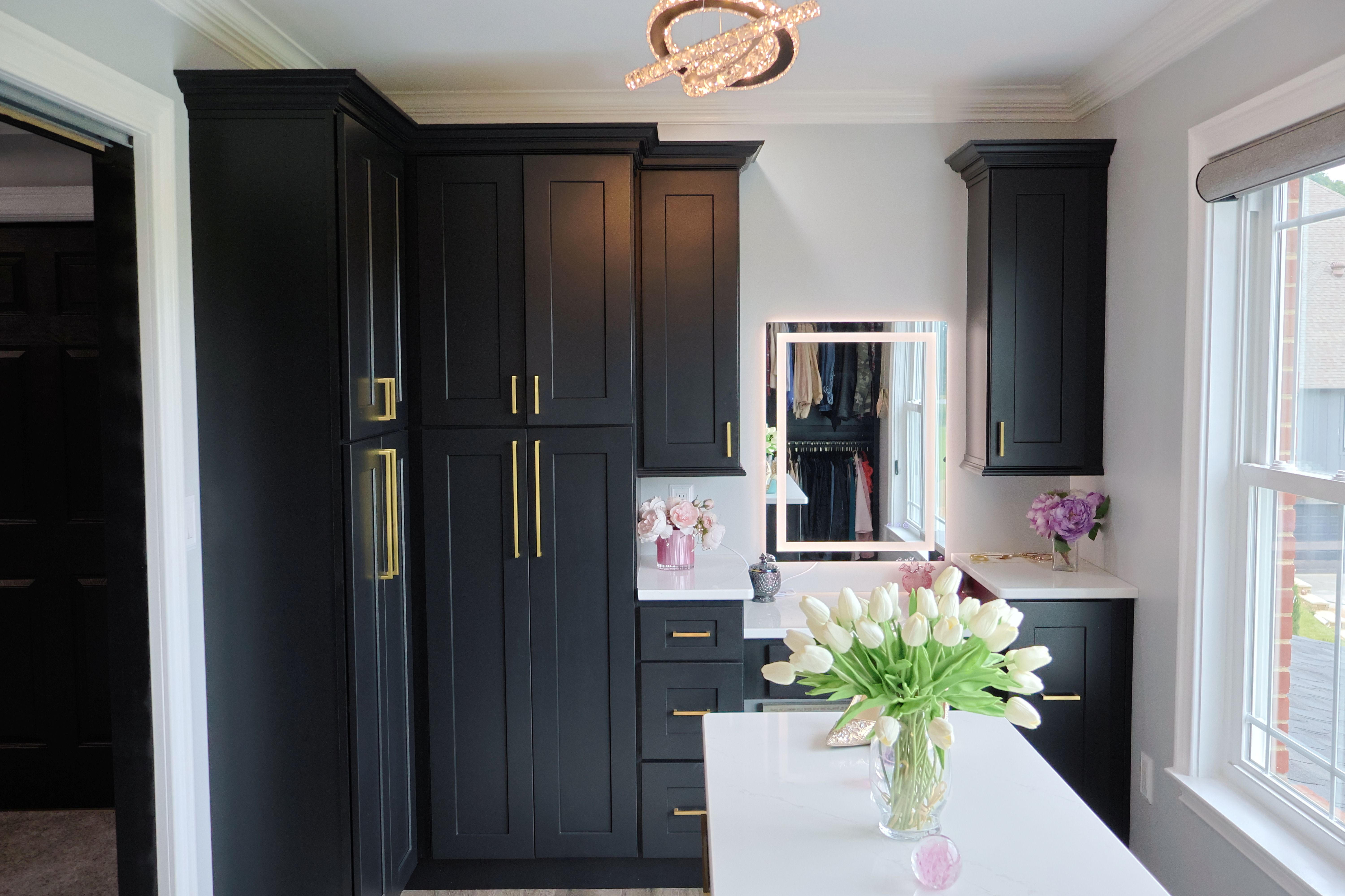 A modern closet with black cabinets featuring gold handles, white countertops, a lighted mirror, and vases with white tulips and purple flowers, next to a window.