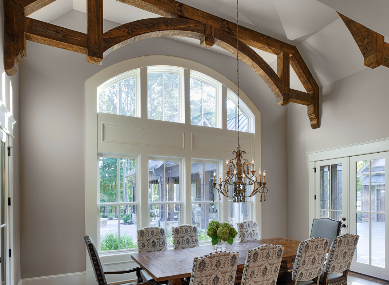 Elegant dining room with large arched windows letting in natural light, a wooden table surrounded by patterned upholstered chairs, and a decorative chandelier hanging from ceiling beams.