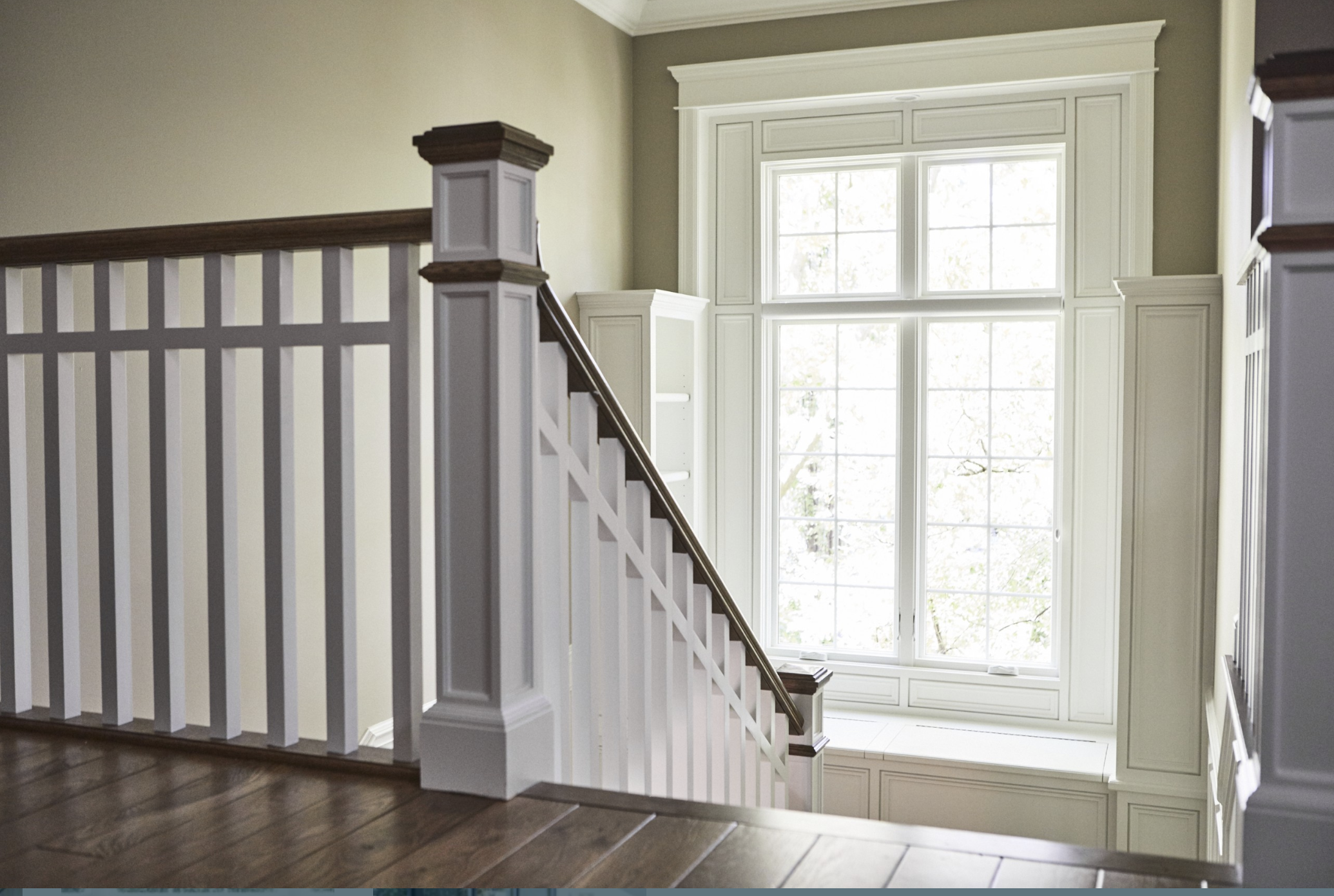 Interior stairway with white wooden railing and dark wood handrail, leading down to a space in front of a large multi-pane window with white trim, flanked by built-in white cabinetry beneath the window.
