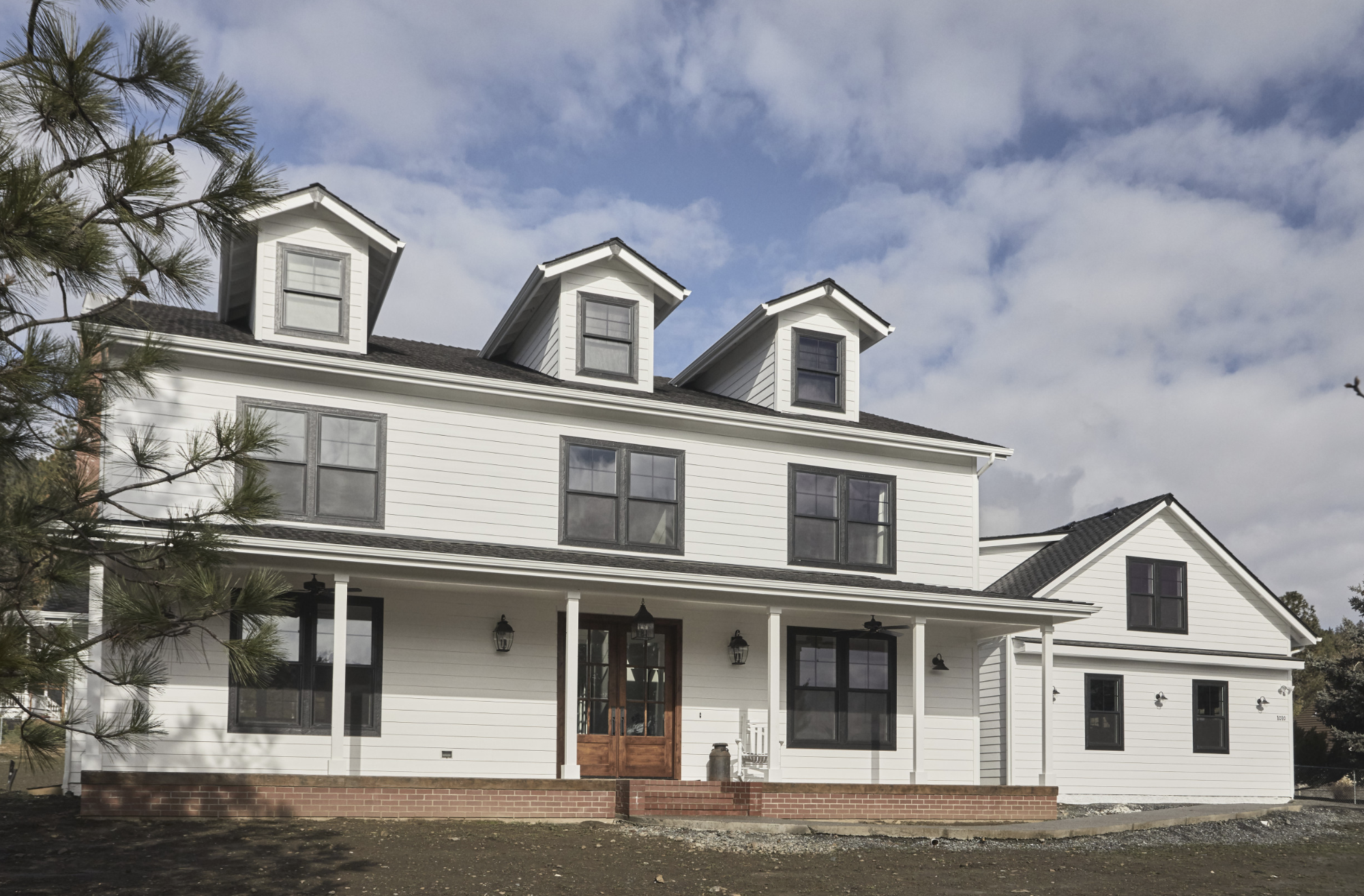 Large white two-story house with three dormer windows on the roof, a brick foundation and porch with wooden double doors and black-framed windows, under a partly cloudy sky.