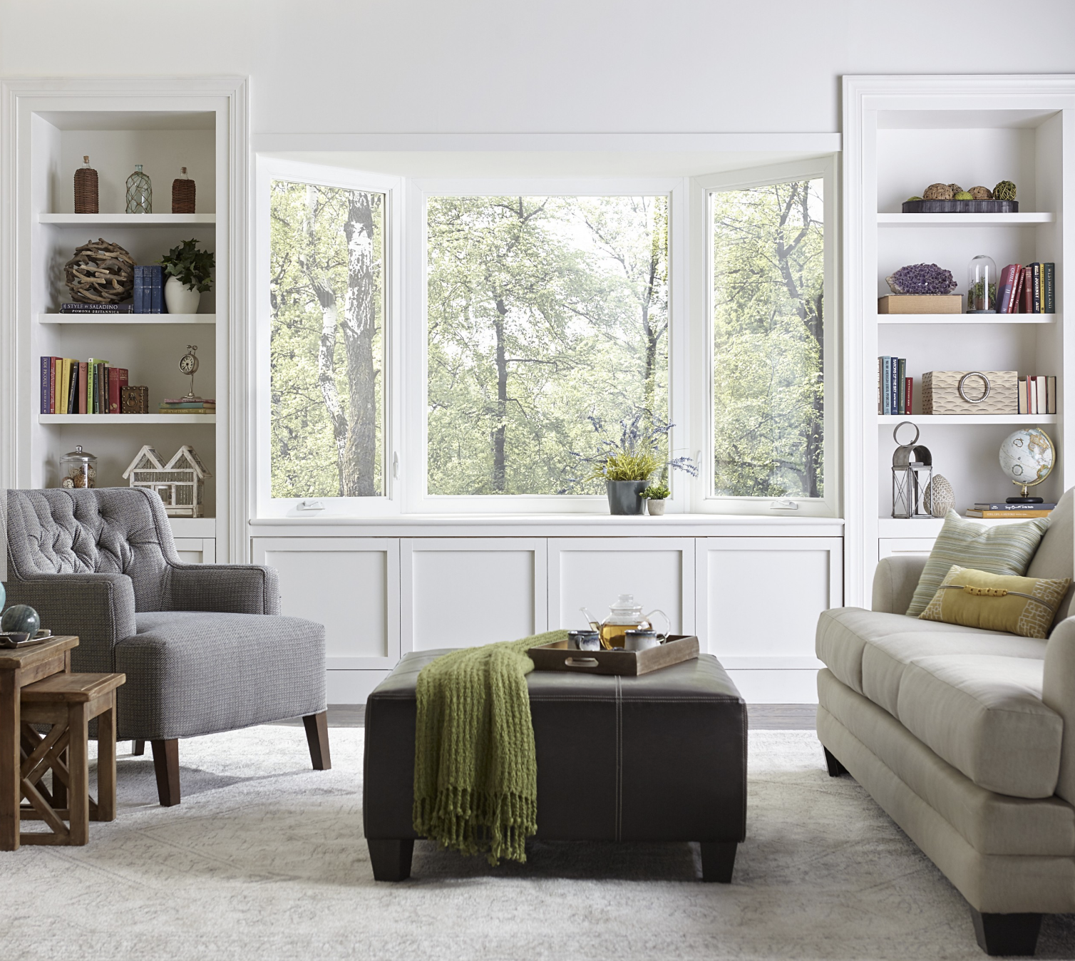 Bright living room with large bay window showing green trees outside, gray armchair on the left, beige sofa with pillows on the right, dark leather ottoman with a green throw and tea set in the center, and built-in white shelves filled with books and decorative objects on each side of the window.