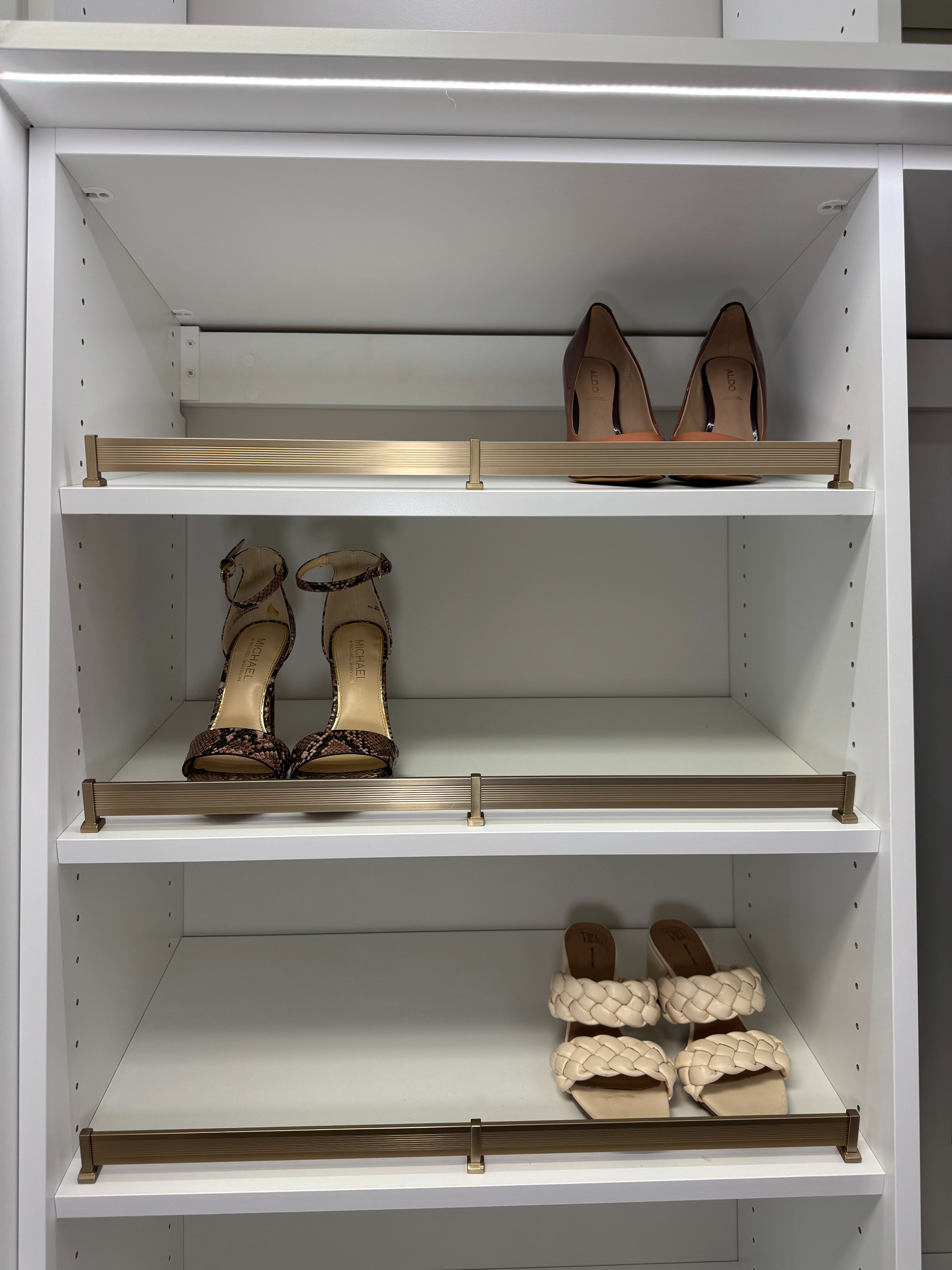 Three pairs of women's shoes arranged on white shelves with gold-colored guard rails: top shelf has brown Aldo heels, middle shelf holds snake-patterned Michael Kors ankle strap heels, and bottom shelf features beige braided open-toe sandals.