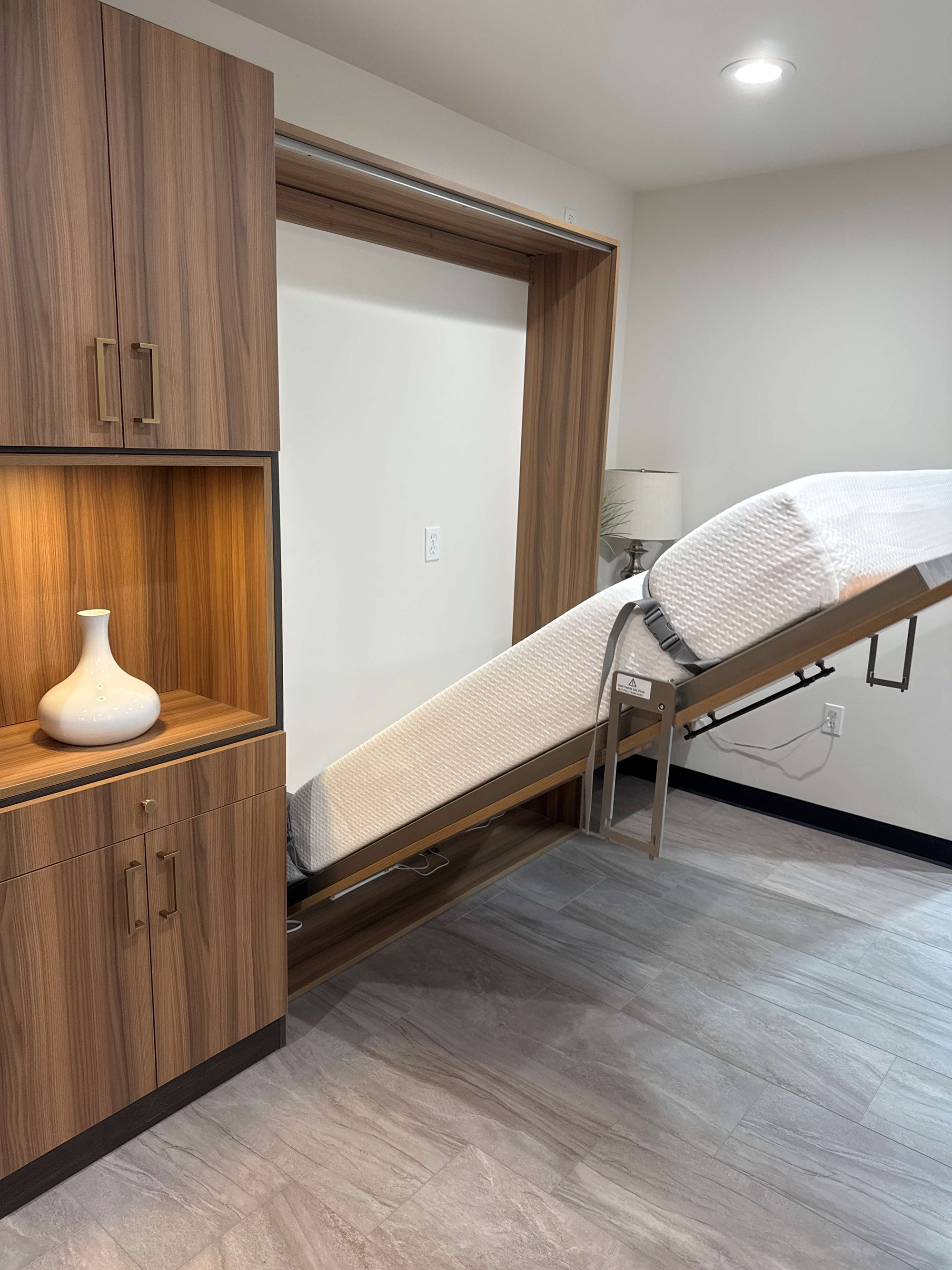 A modern Murphy bed partially folded down from a wooden wall cabinet in a room with gray tiled floor, white walls, and a small white vase on a shelf to the left.