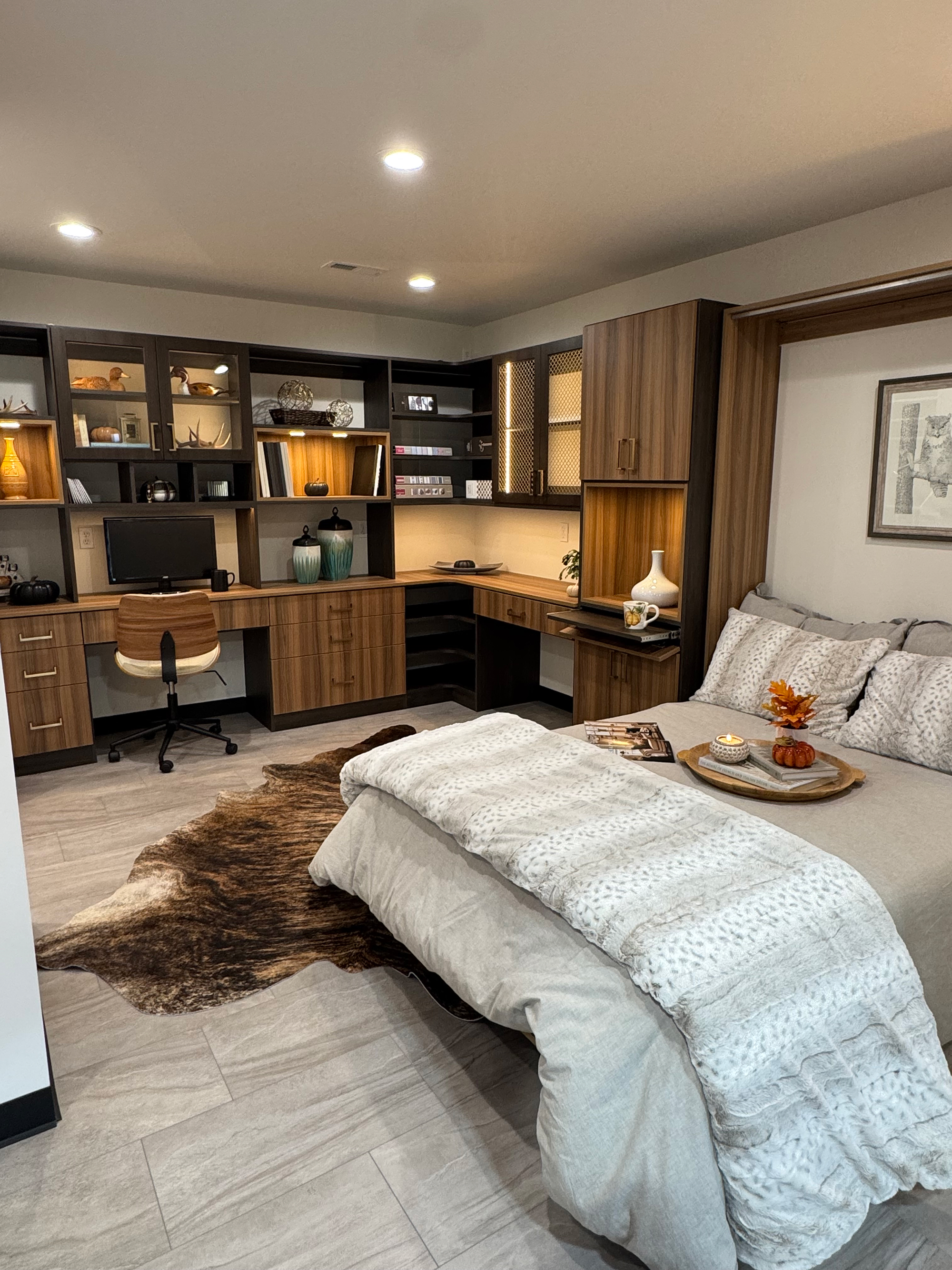 Modern bedroom with a beige bed covered in a textured white throw blanket, a tray with a small pumpkin and candle on the bed, a brown and white office chair at a corner wooden desk with shelves holding decorative items like vases, books, and sculptures, and a large animal print rug on light gray tile flooring.