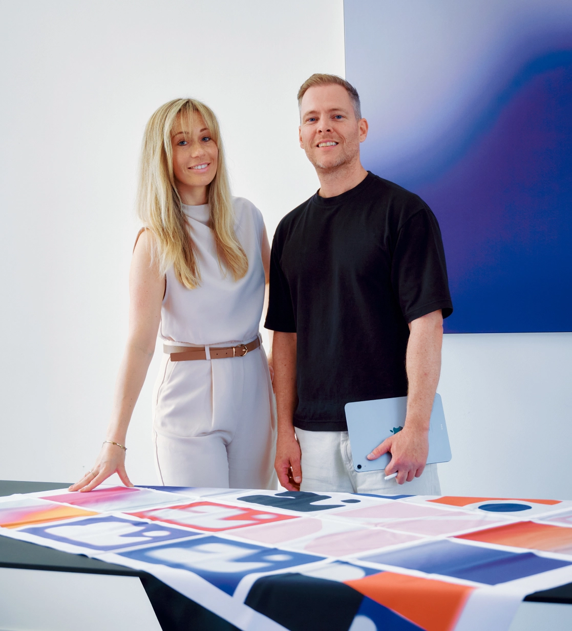A woman in a light sleeveless outfit and a man in a black t-shirt holding a tablet stand beside a table covered with a colorful patterned fabric, smiling at the camera.