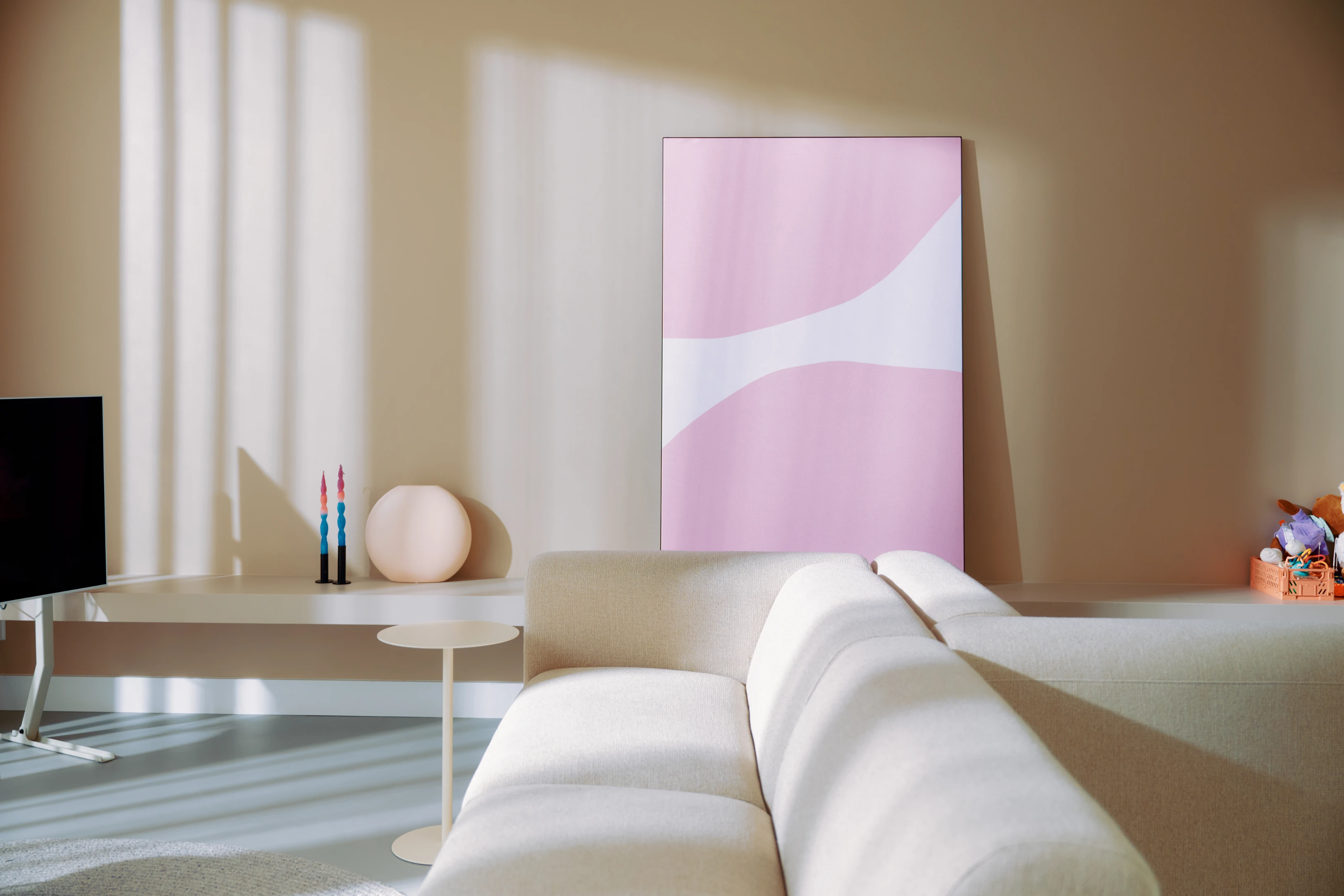 Modern living room with beige sofa facing a TV, a small round side table, pastel abstract pink and white artwork on the wall, and decorative candles and items on a white console table.