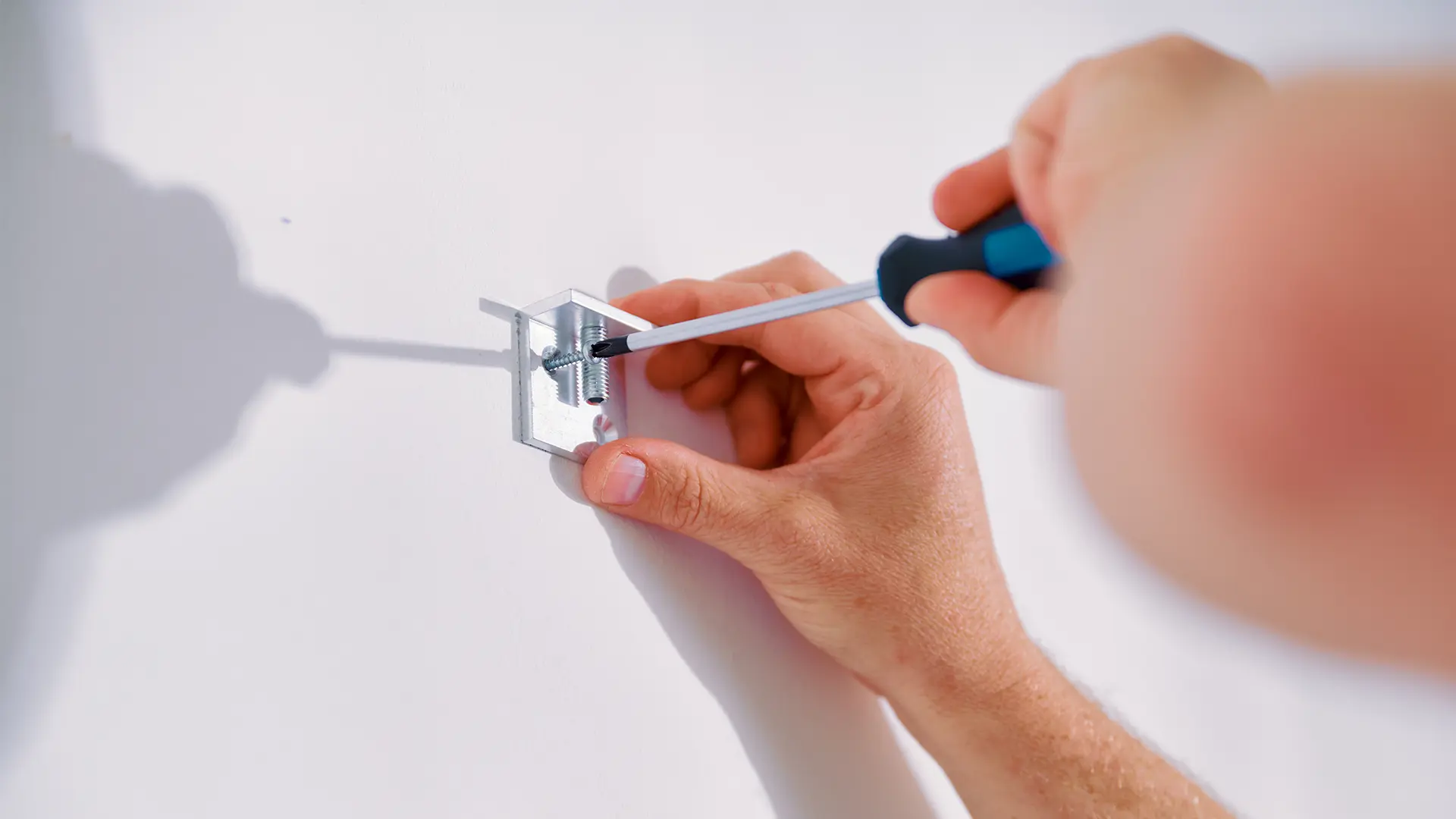 Hand using a screwdriver to attach a metal bracket to a white wall.