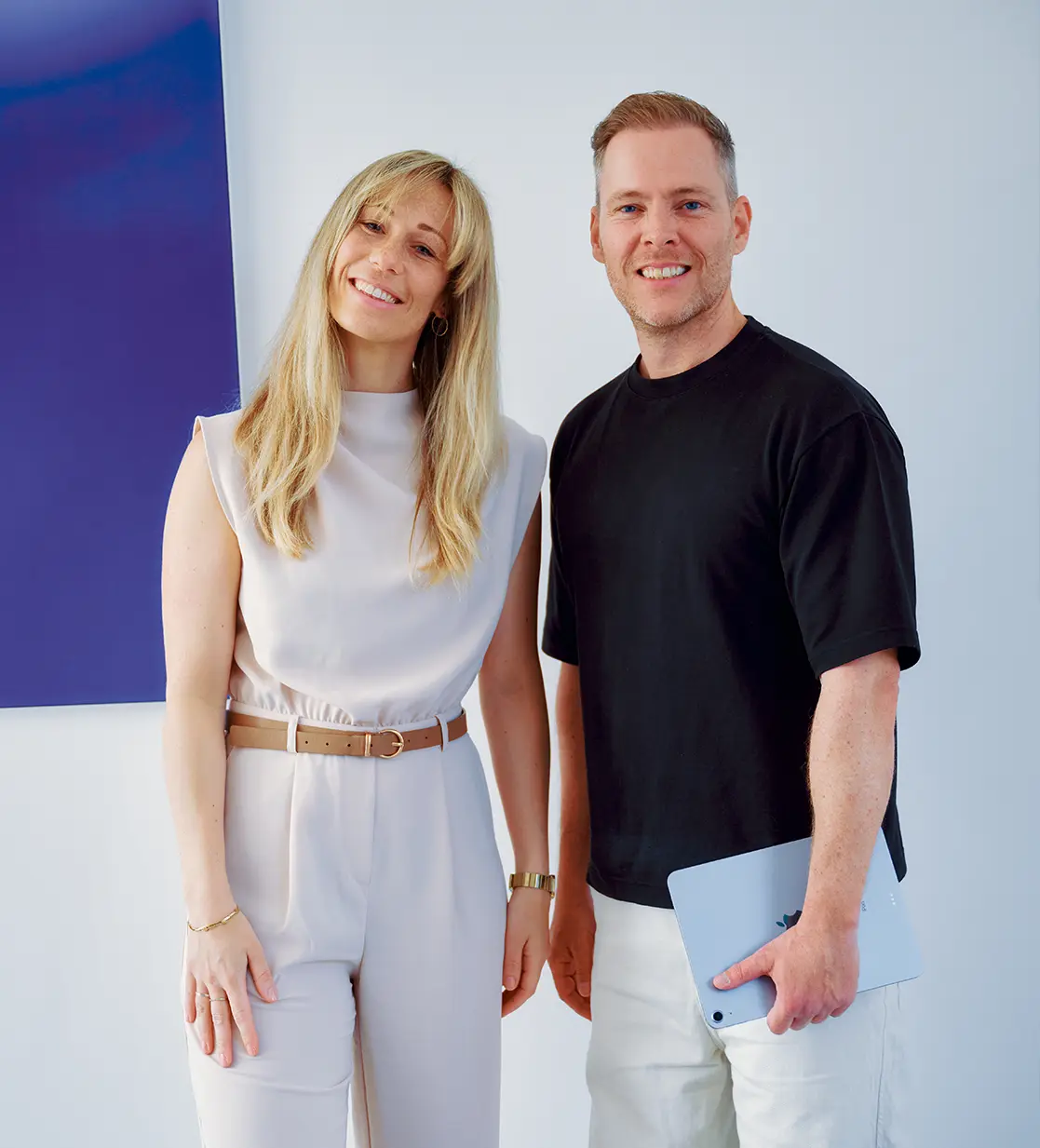 Smiling woman with long blonde hair wearing a white outfit with a tan belt standing next to a smiling man in a black shirt holding a tablet.