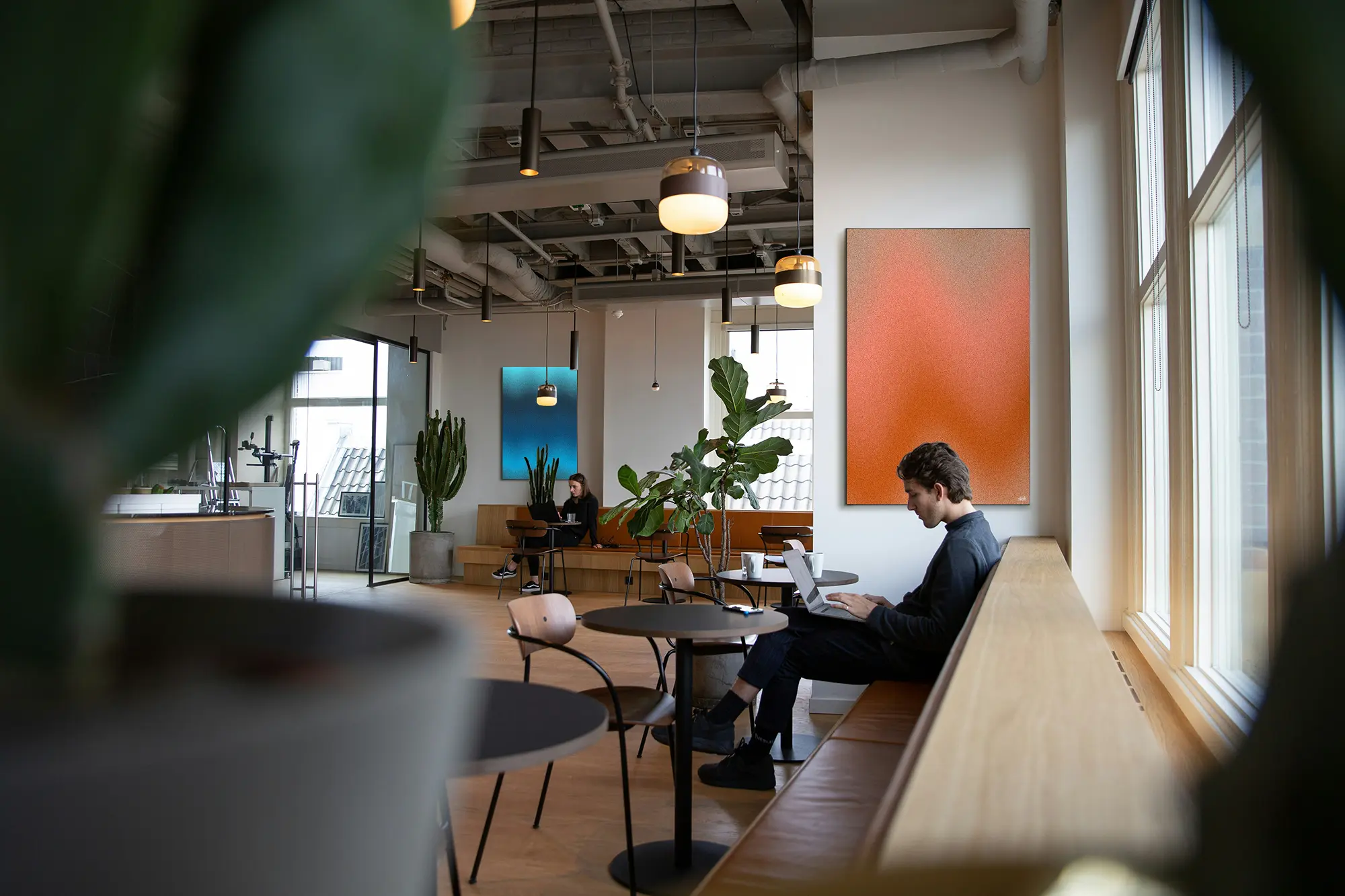 Two people working on laptops in a modern, plant-filled office lounge with pendant lights and colorful abstract paintings.