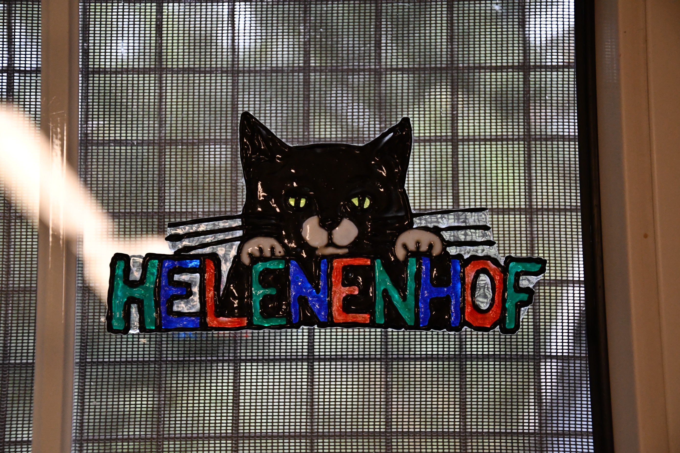 Bunte Glasdekoration eines schwarzen Katzenkopfs mit grünen Augen, der die Buchstaben HELENENHOF hält.