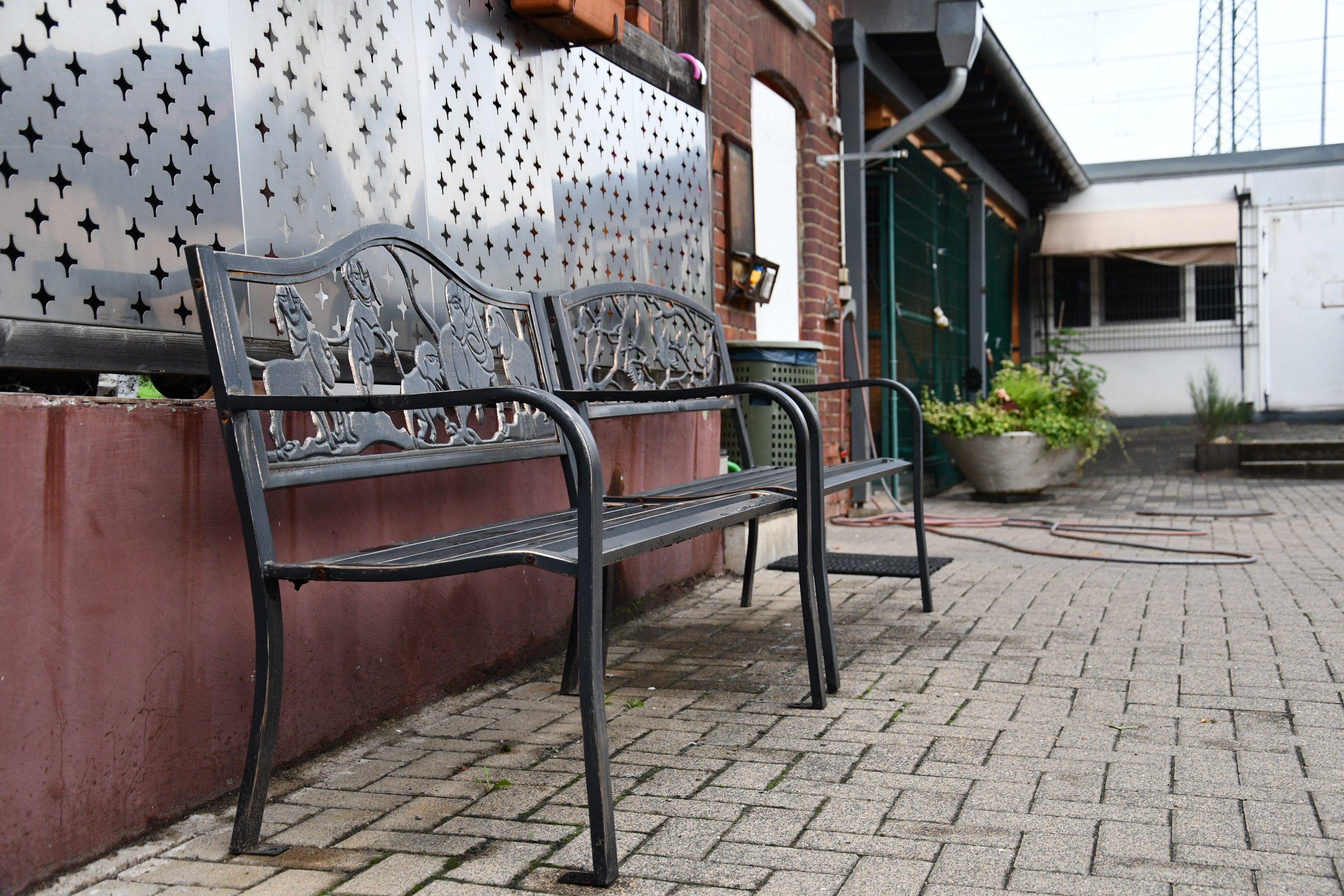 Zwei metallene Gartenbänke mit Verzierungen stehen auf einem gepflasterten Hof neben einem roten Mauersockel und Pflanzen.