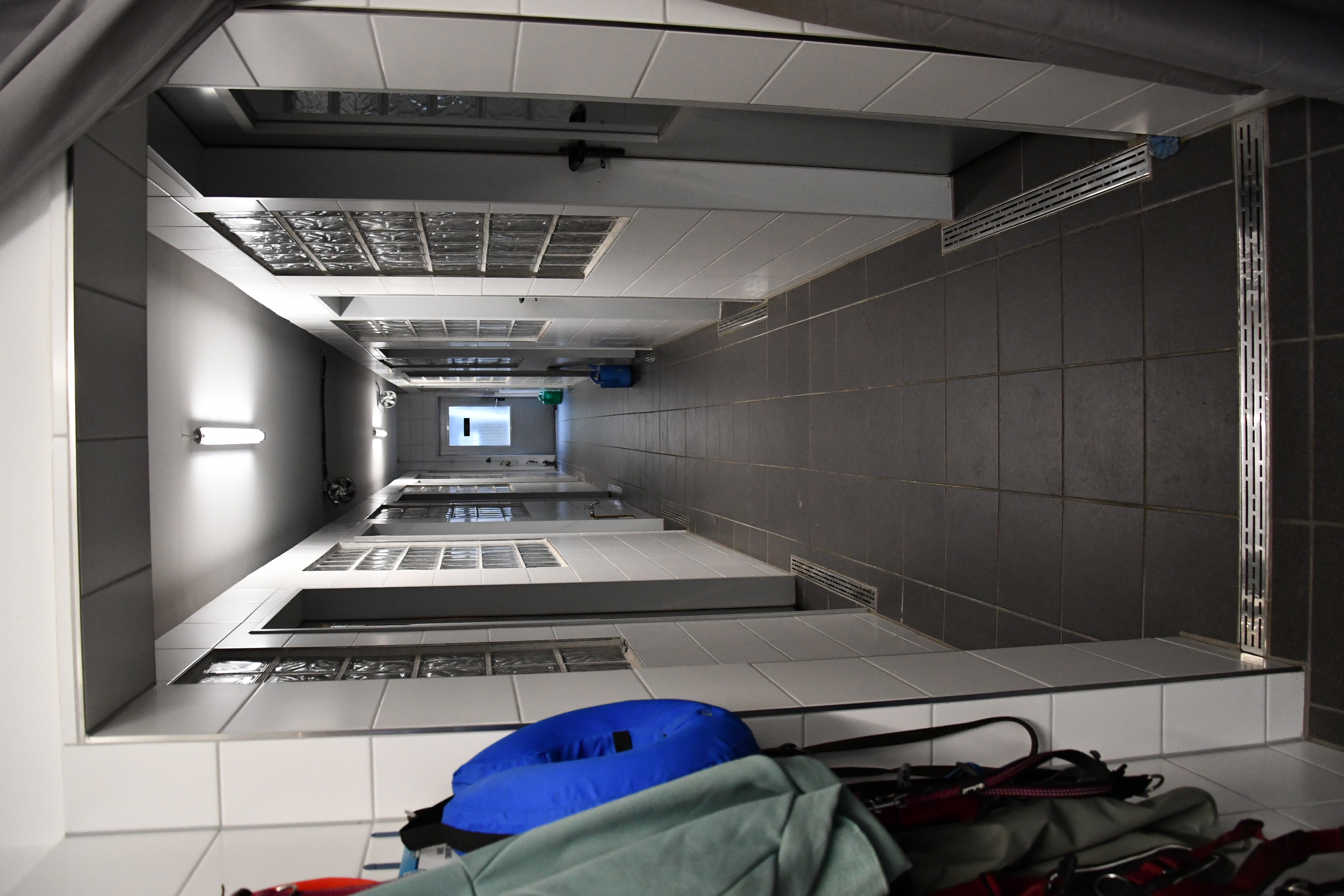 Flur mit dunklem Fliesenboden und weißen Wänden, mehrere Türen und Glasbausteine, beleuchtet von Deckenlampen.