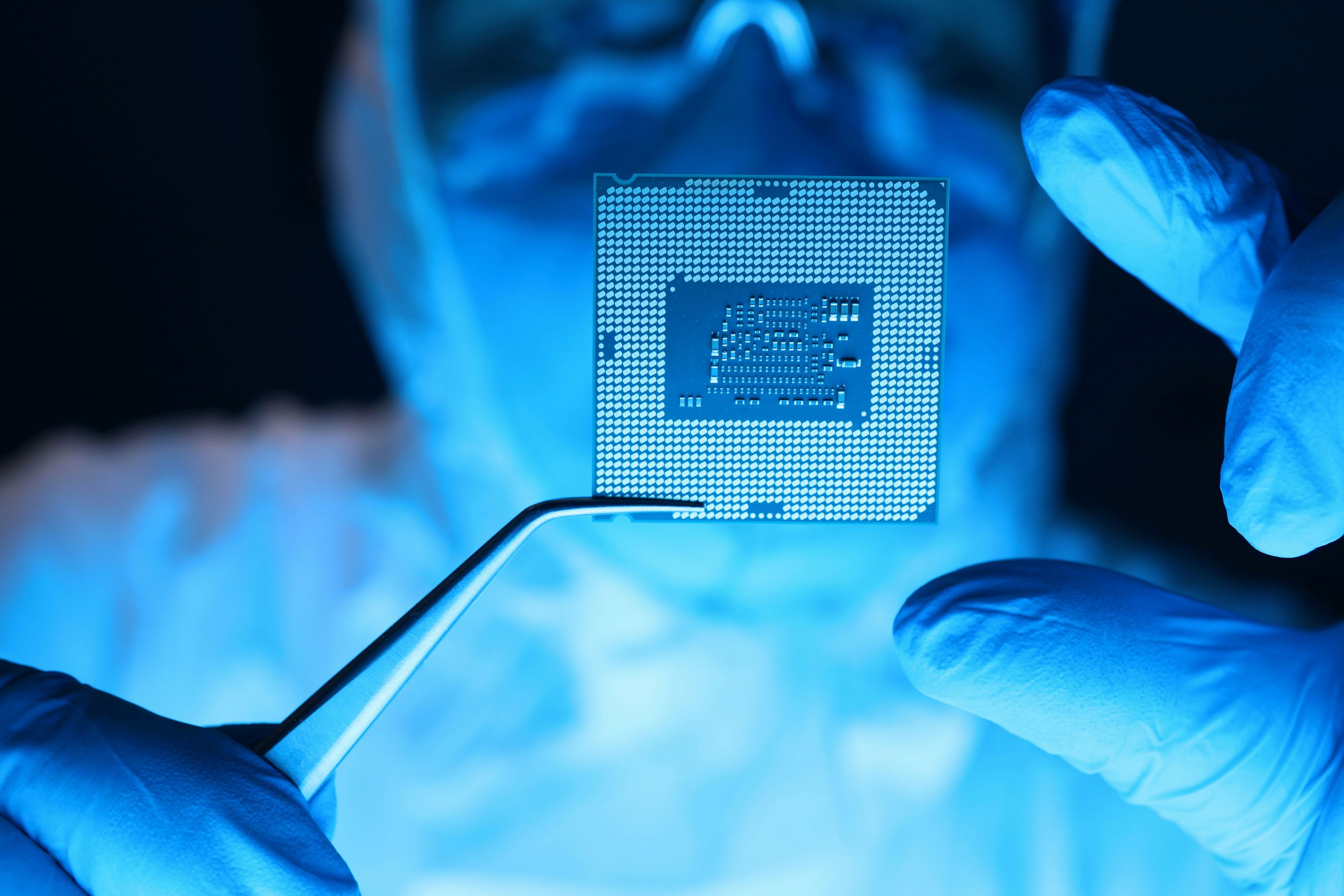 Technician in cleanroom gloves holding a microchip with precision tweezers under blue lighting, representing high-tech electronics manufacturing and semiconductor inspection.

