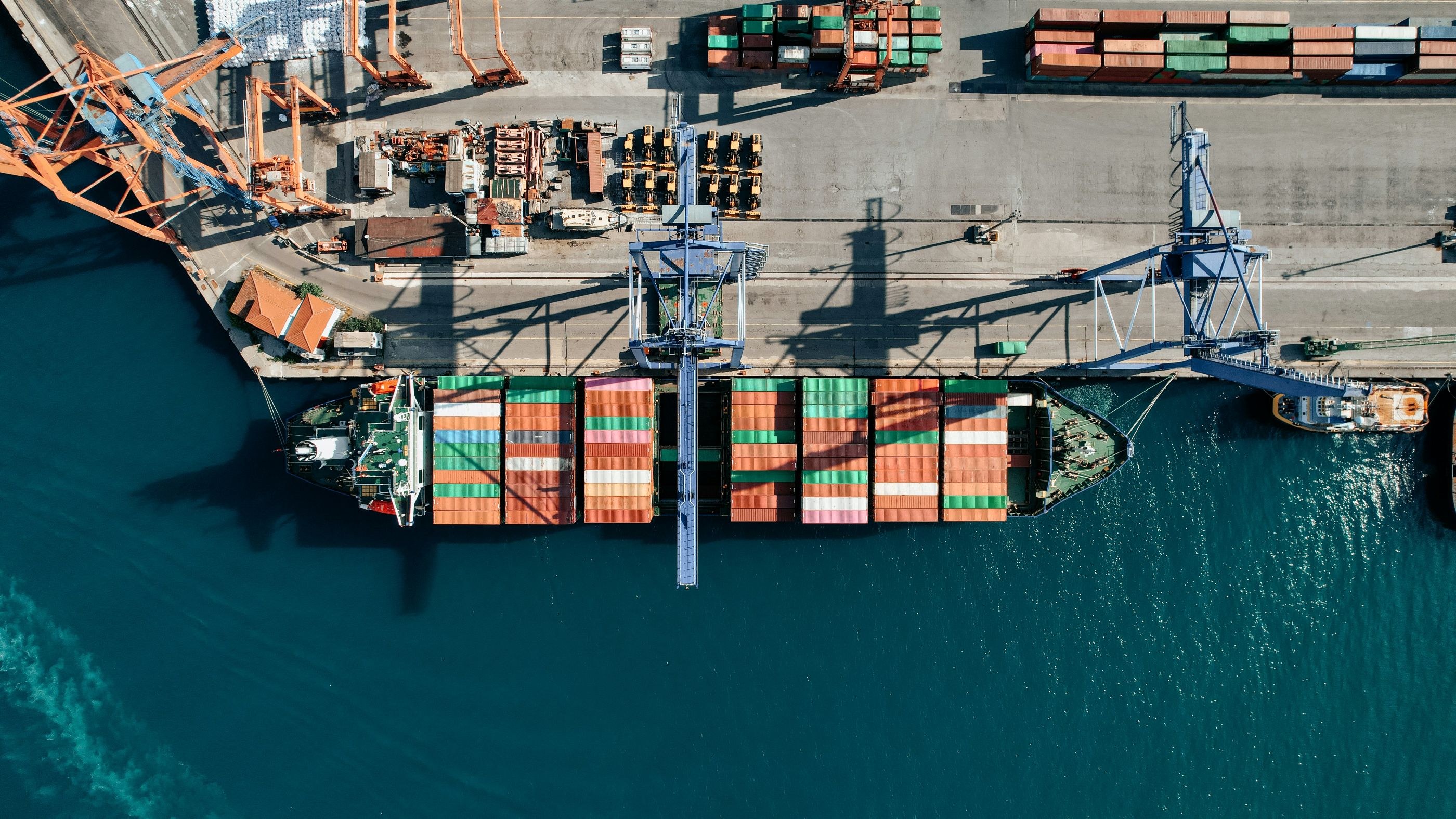 Aerial view of a cargo ship docked at a port terminal with colorful shipping containers being loaded by cranes, representing global trade and logistics operations.

